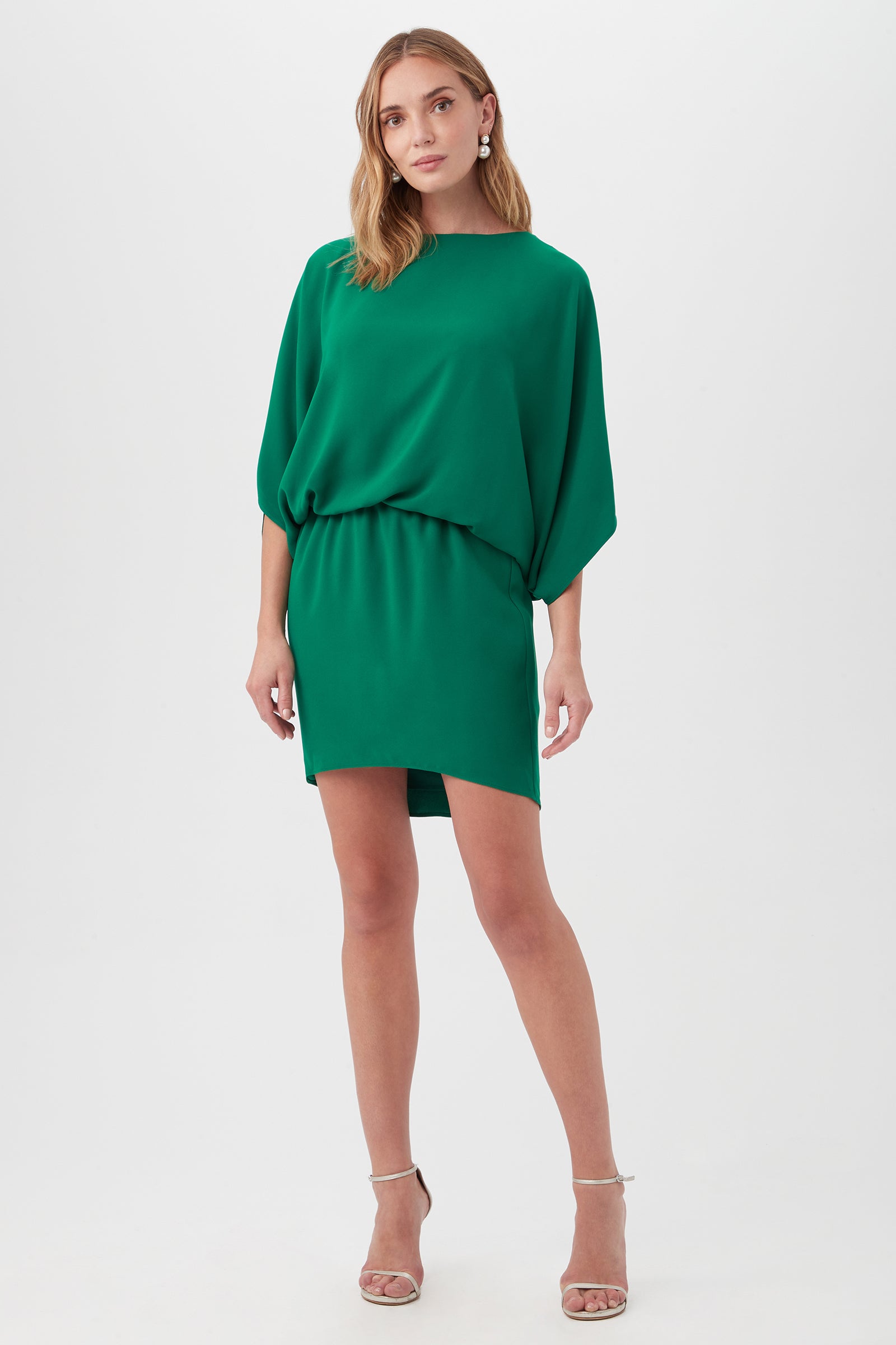 A woman poses against a plain white background in the Manhattan Dress—a loose-fitting, short green dress with wide sleeves and an elastic waist—paired with strappy nude high-heeled sandals. Her light brown hair is styled in loose waves.