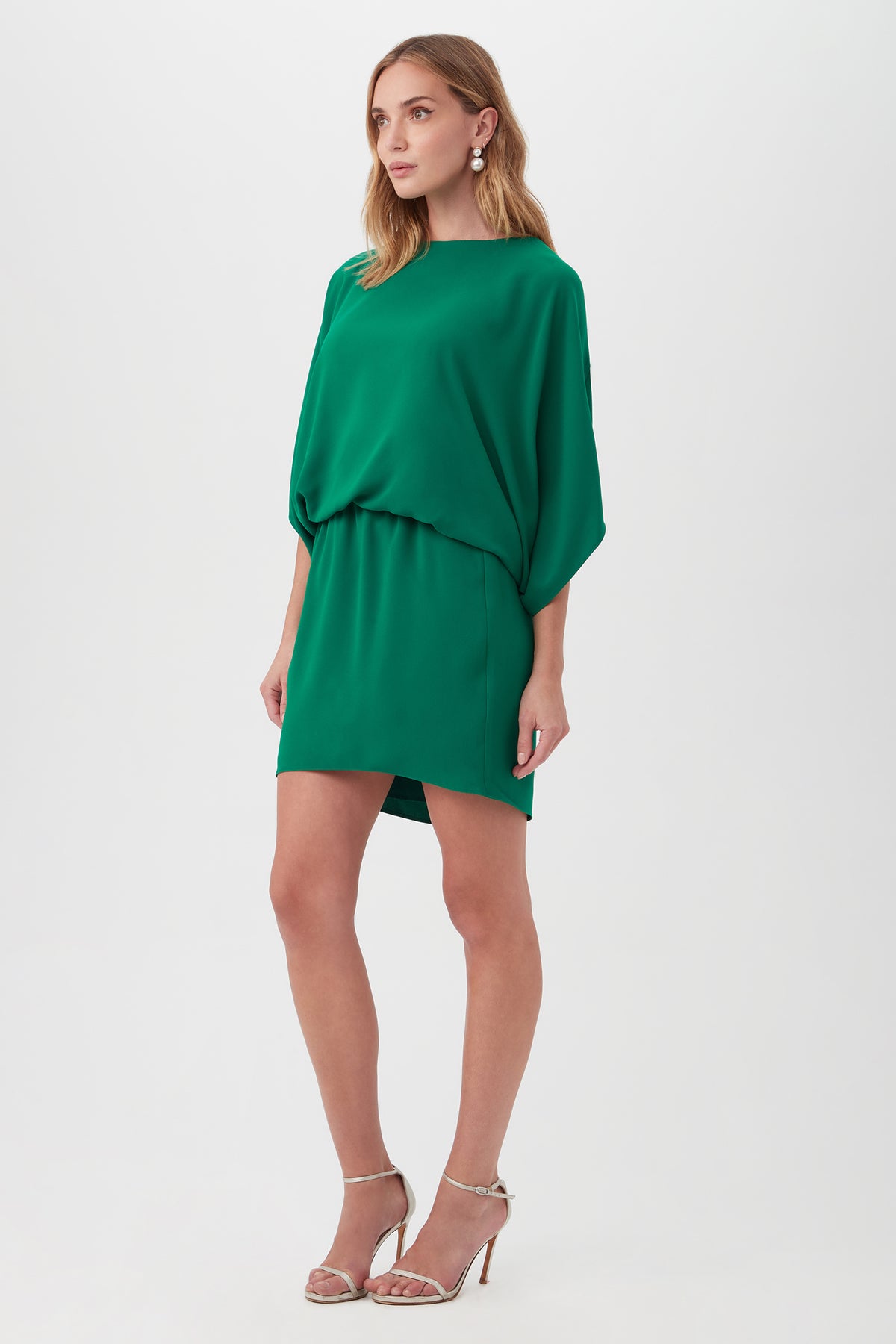 A woman wears the Manhattan Dress—a loose-fitting, short green dress with elbow-length sleeves—paired with silver high-heeled sandals, standing against a plain white background.