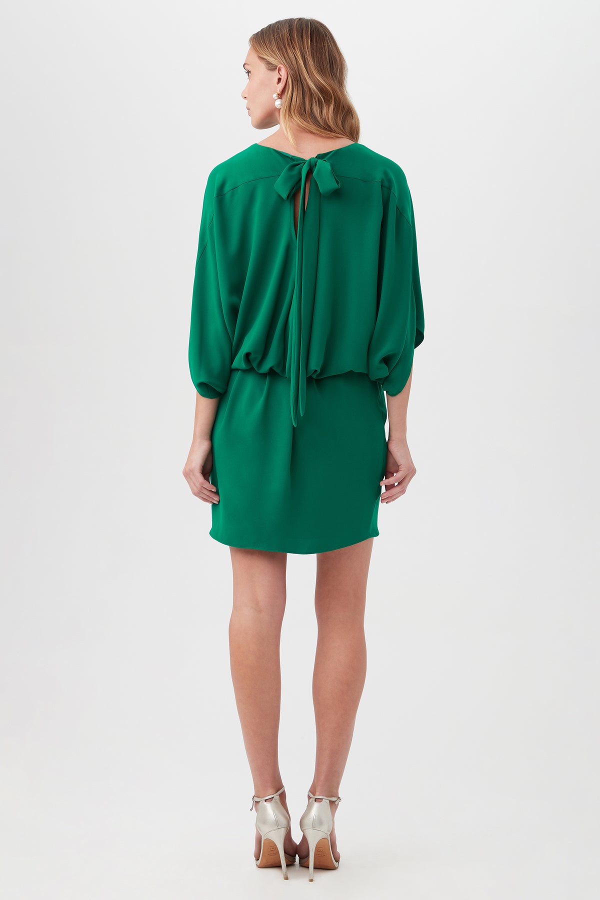 A woman faces away in the Manhattan Dress, a loose-fitting, knee-length green dress with a tied neck bow. She has wavy light brown hair and wears white high-heeled sandals against a plain white background.