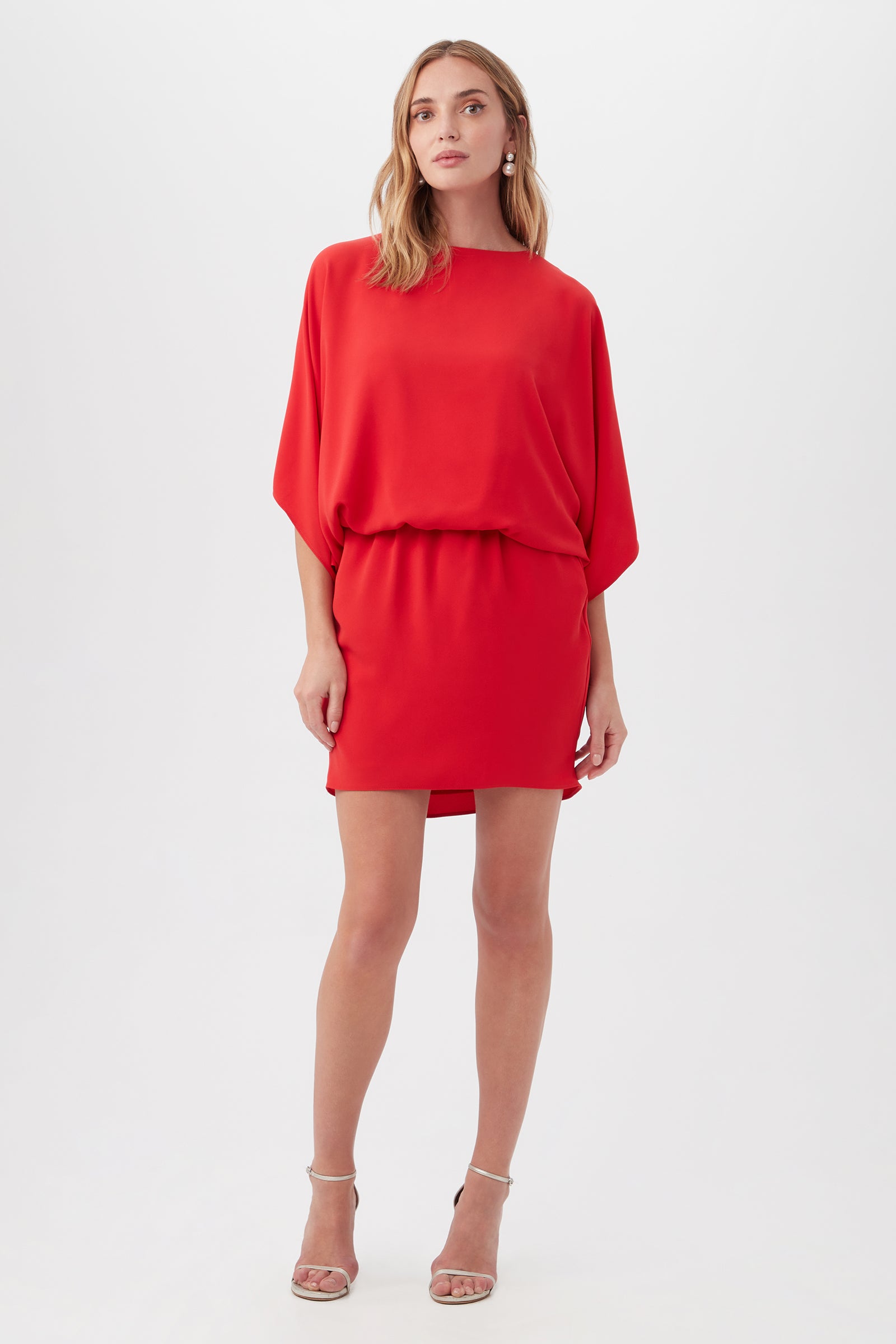 A woman poses against a white background in the Manhattan Dress in Reina Red, featuring a loose fit, batwing sleeves, and an elastic waist, styled with strappy high-heeled sandals.