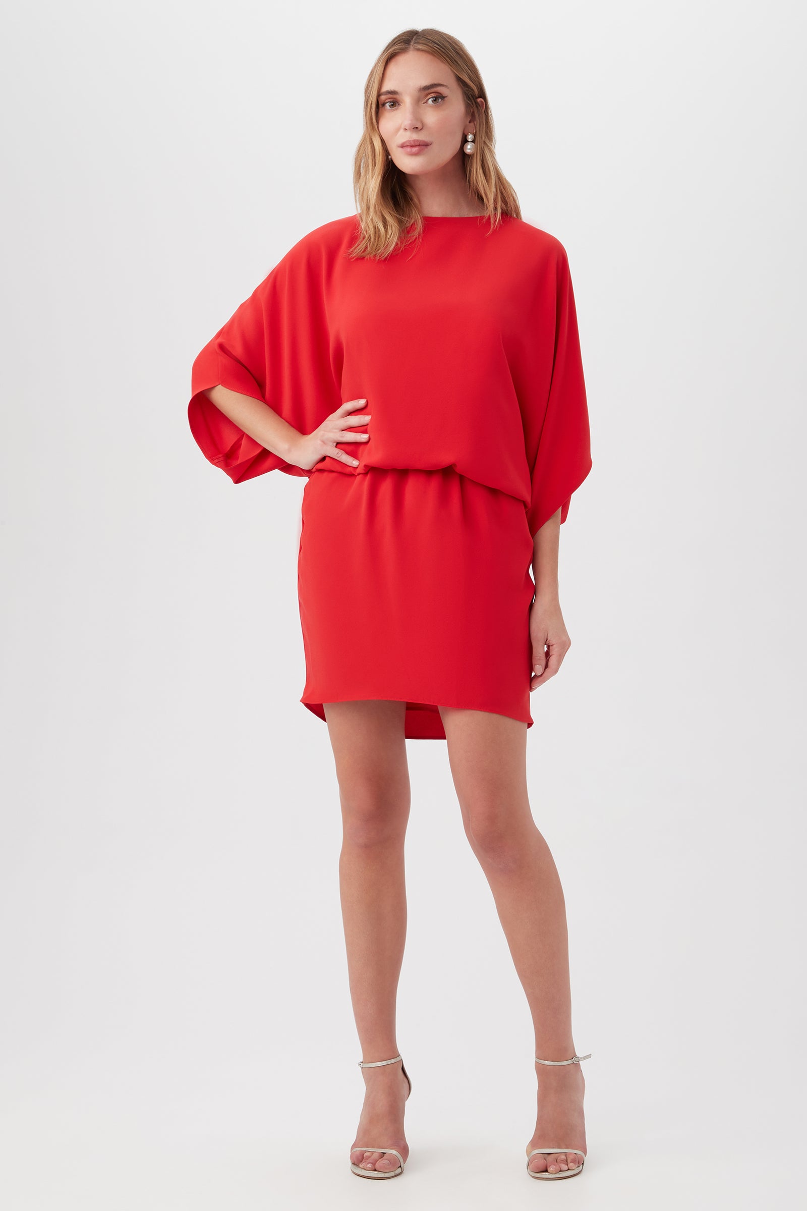 A woman stands confidently in the Manhattan Dress—a vibrant red, loose-fit, short-sleeve piece with a relaxed waist and short hem—paired with strappy nude high-heeled sandals, set against a plain light background.