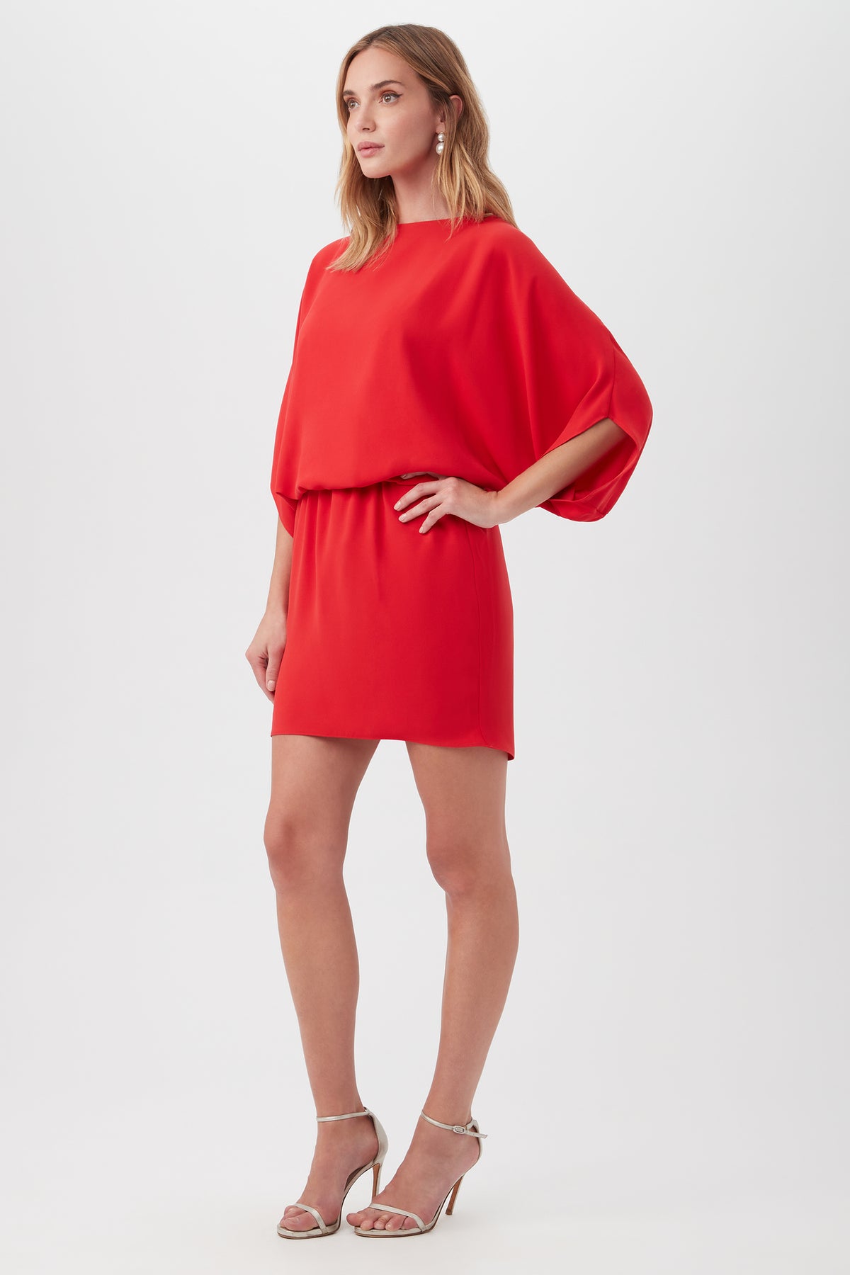 A woman models the Manhattan Dress—a red, loose-fit, short-sleeve dress with a cinched waist and short skirt—paired with silver high-heeled sandals against a plain white background.