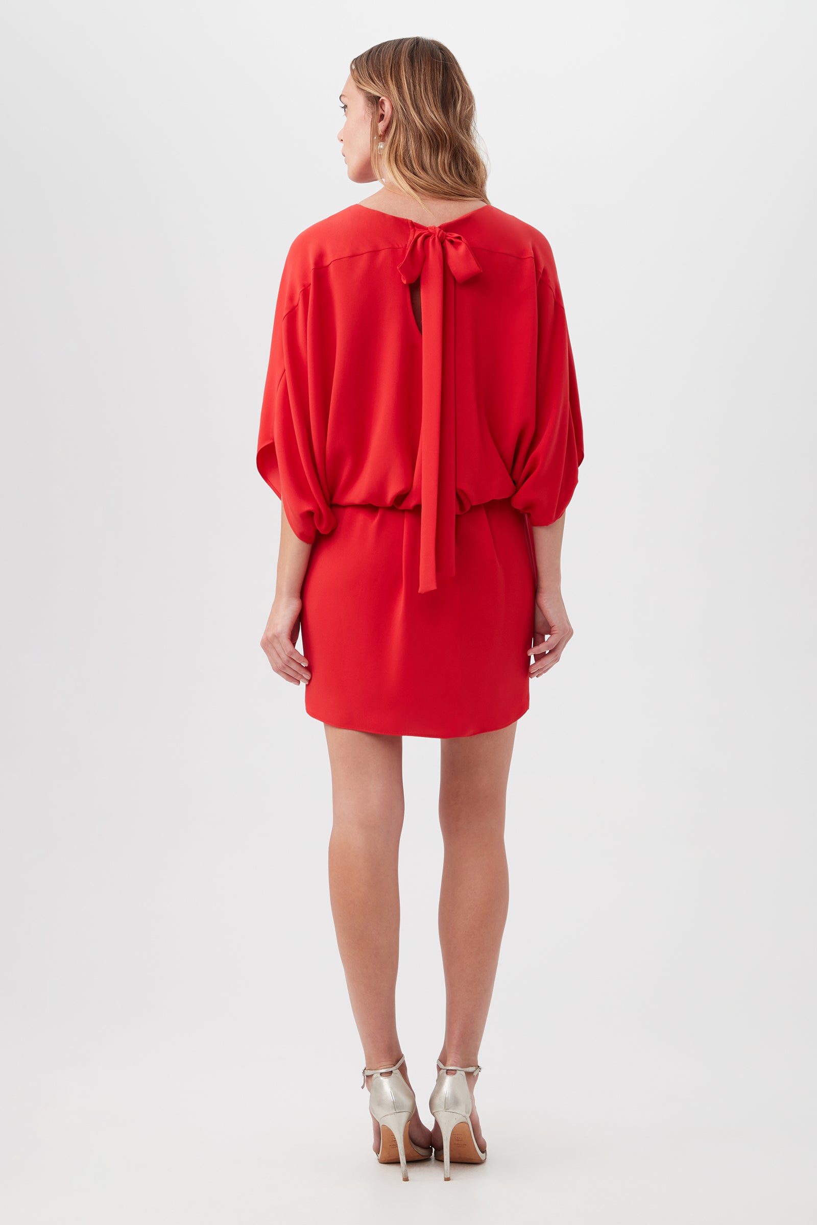 A woman faces away, wearing the Manhattan Dress—a short red style with loose sleeves, a back neck bow tie—and silver high-heeled sandals against a plain white background.