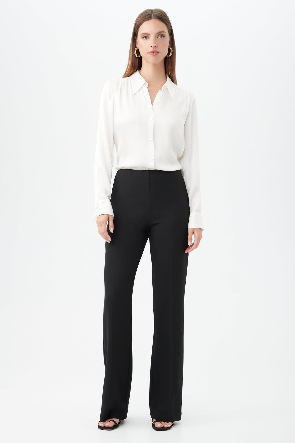 A woman stands against a plain white background wearing the Cassavetes Long-Sleeve Stretch Silk Button-Up Shirt, black high-waisted wide-leg pants, black open-toe heels, and gold hoop earrings. She has straight, shoulder-length hair.