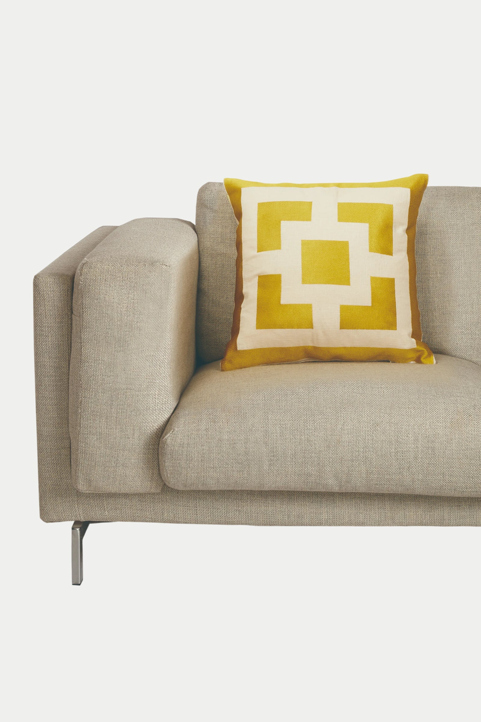 A modern beige sofa with square armrests features the PALM SPRINGS BLOCK EMB PILLOW 20X20", a yellow and white cushion filled with down feathers, set against a plain white background.