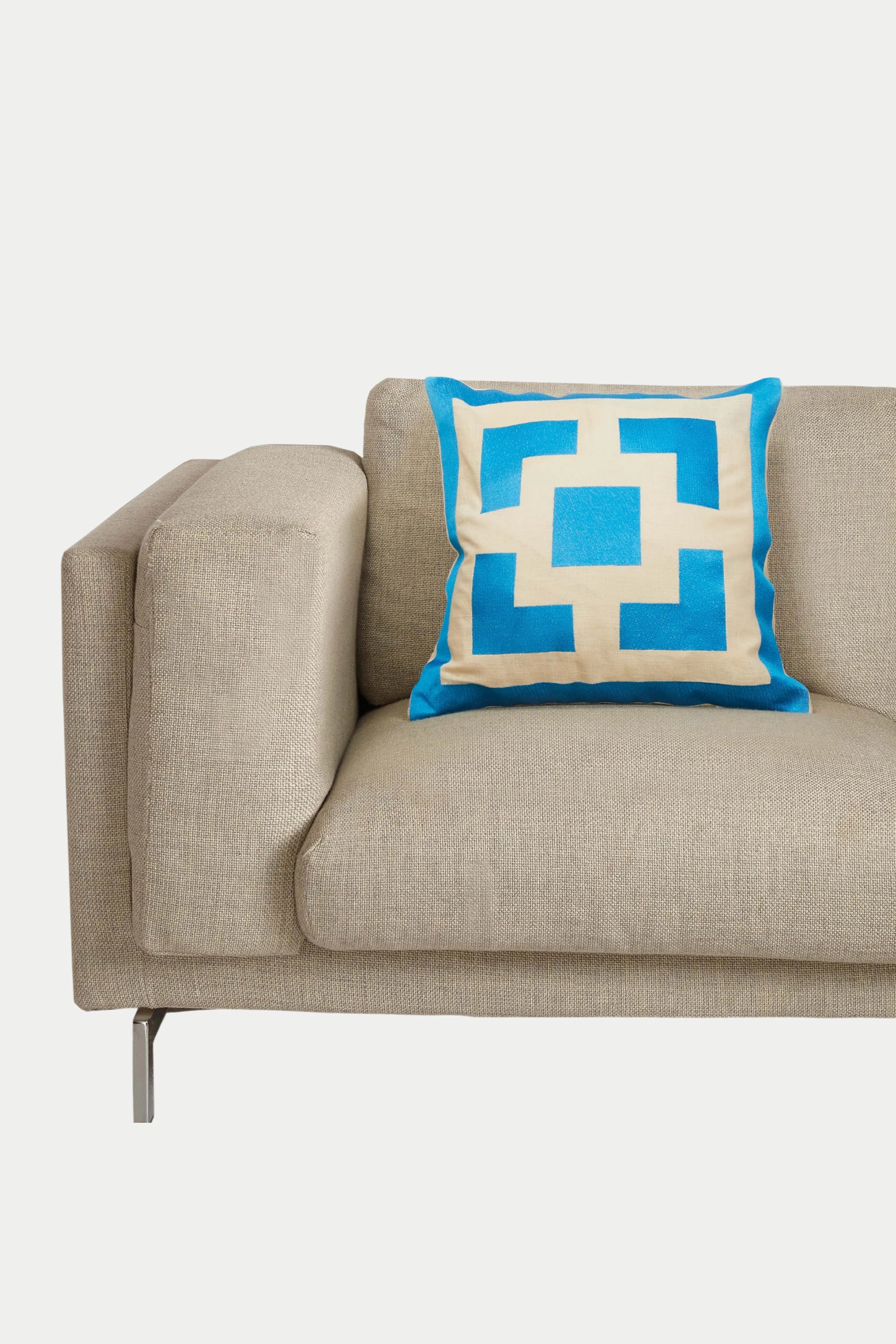 A beige modern sofa with metal legs features the Ps Block Emb Pillow 20X20", which has a blue and cream geometric design and plush down insert, all set against a plain white background.