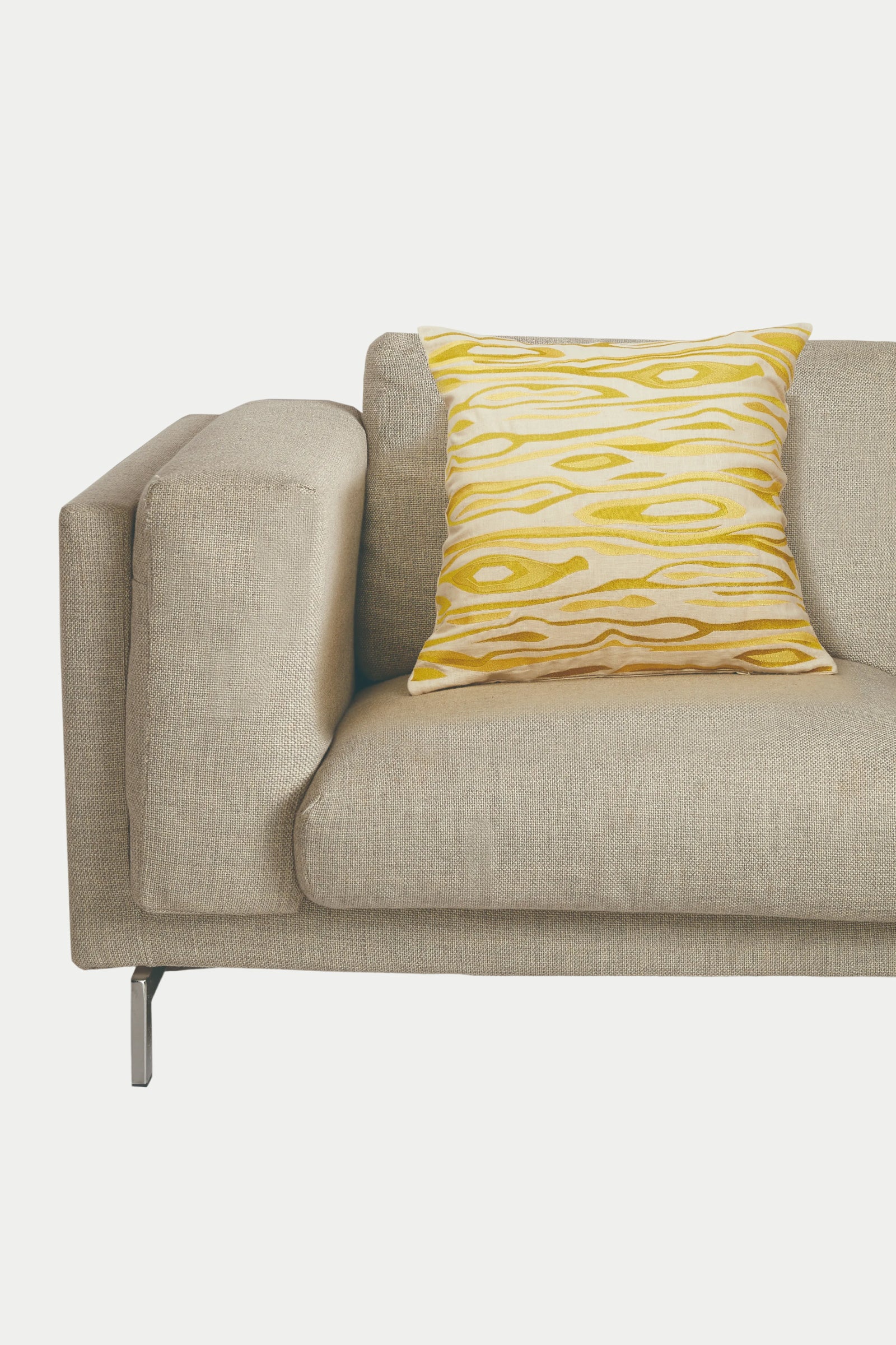 A beige modern sofa with metal legs displays the Monterey Emb Pillow 20x20", featuring a yellow and white pattern and down feather fill, set against a plain white background.