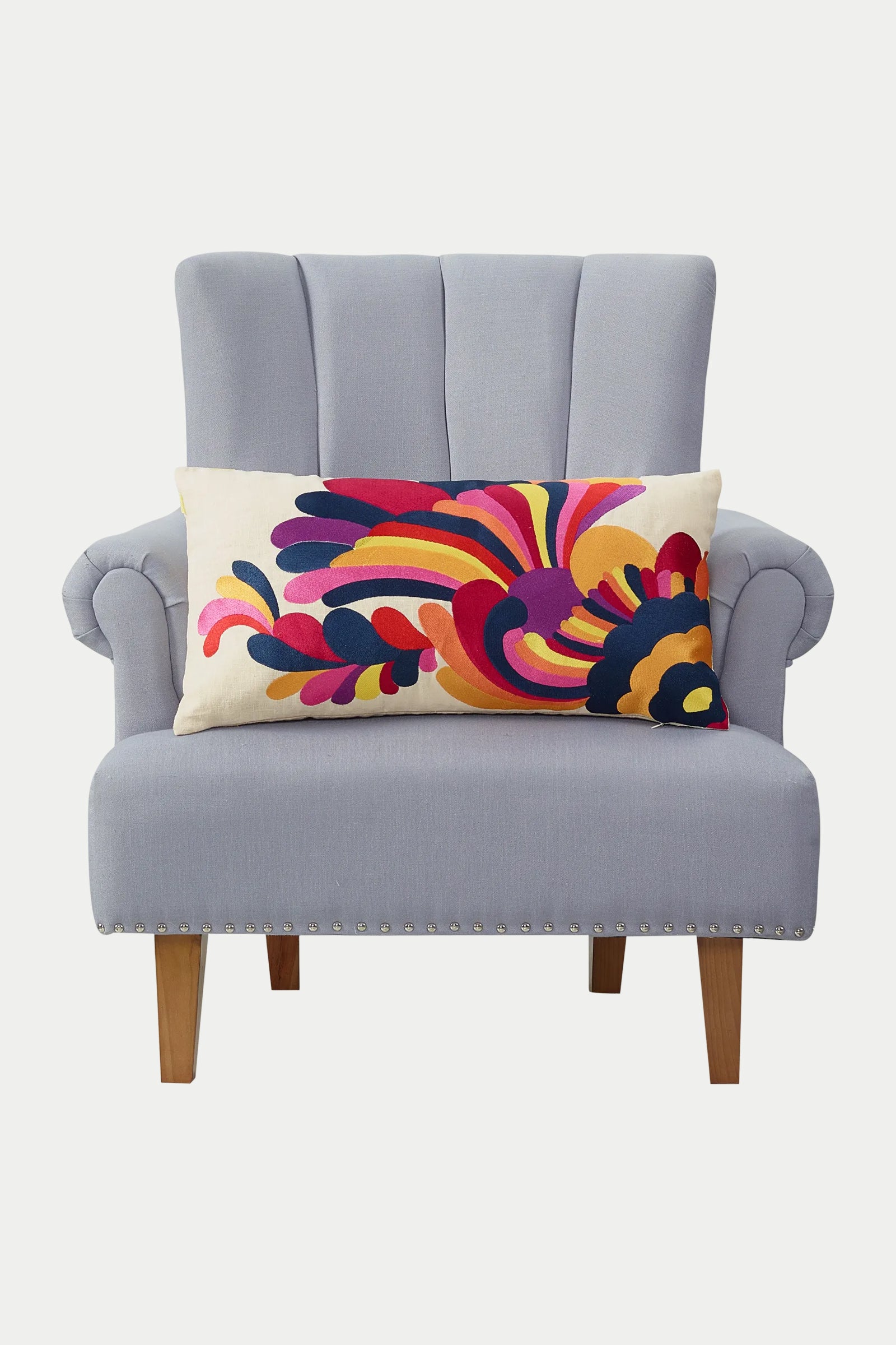 A light gray upholstered armchair with wooden legs features the Escondido Embroidered Throw Pillow in multi, showcasing a colorful abstract pattern in red, pink, yellow, purple, and navy blue.