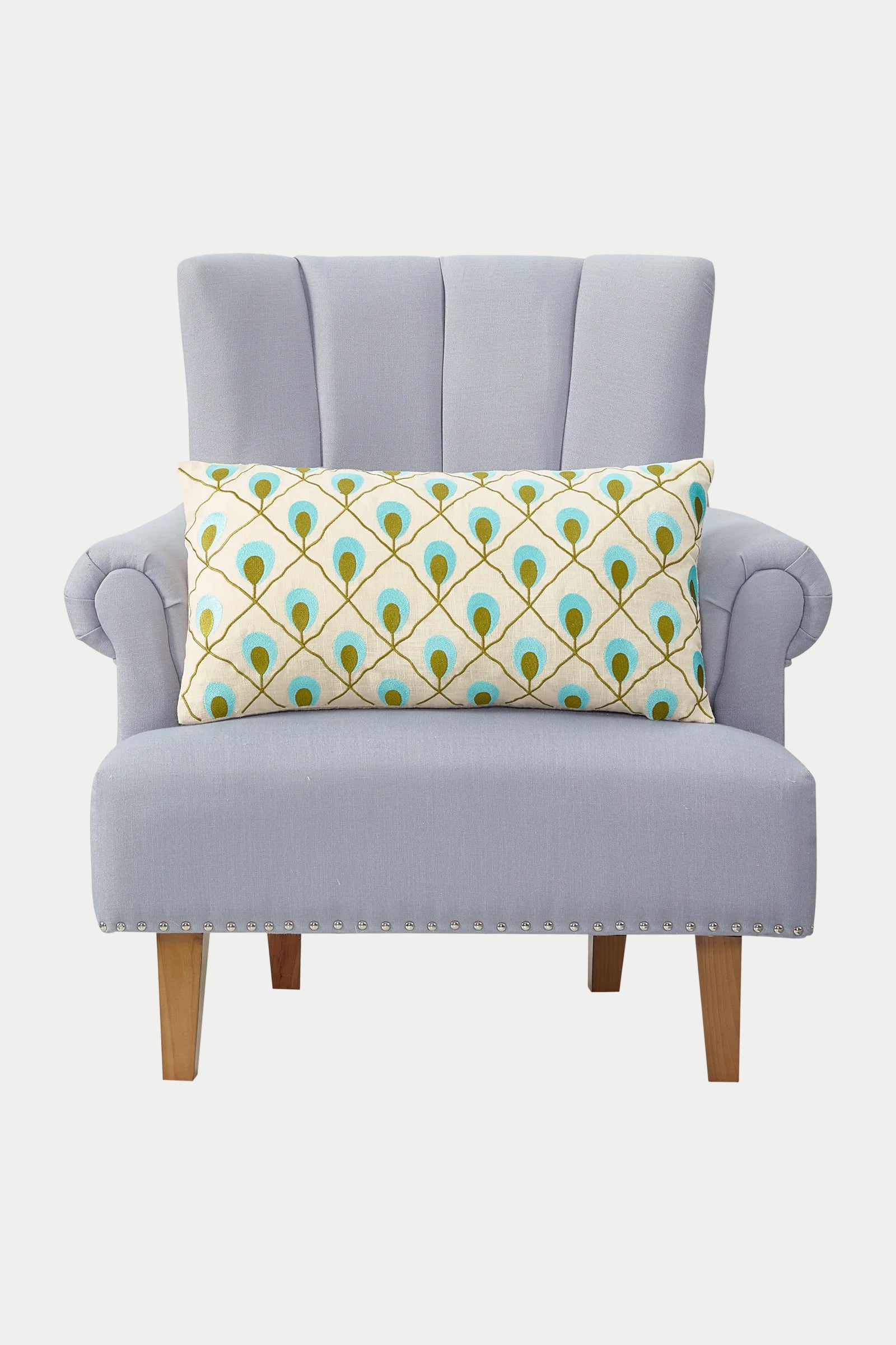 A light gray upholstered armchair with wooden legs, featuring the Roseville Embroidered Throw Pillow in blue with a long rectangular shape and a yellow and turquoise peacock feather pattern.