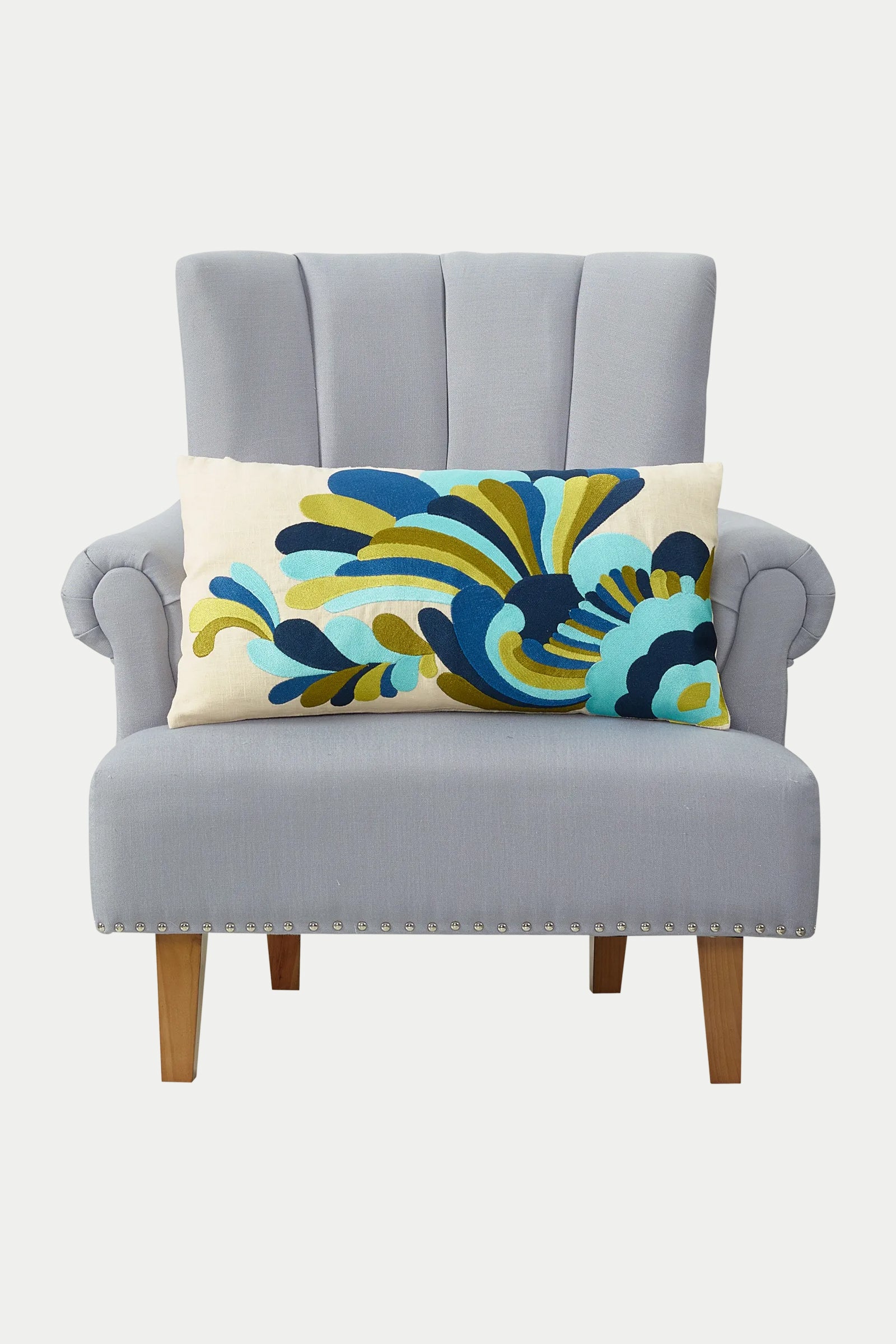 A light gray upholstered armchair with wooden legs, styled with the Escondido Embroidered Throw Pillow in blue, featuring a bold abstract pattern in blue, green, and yellow.