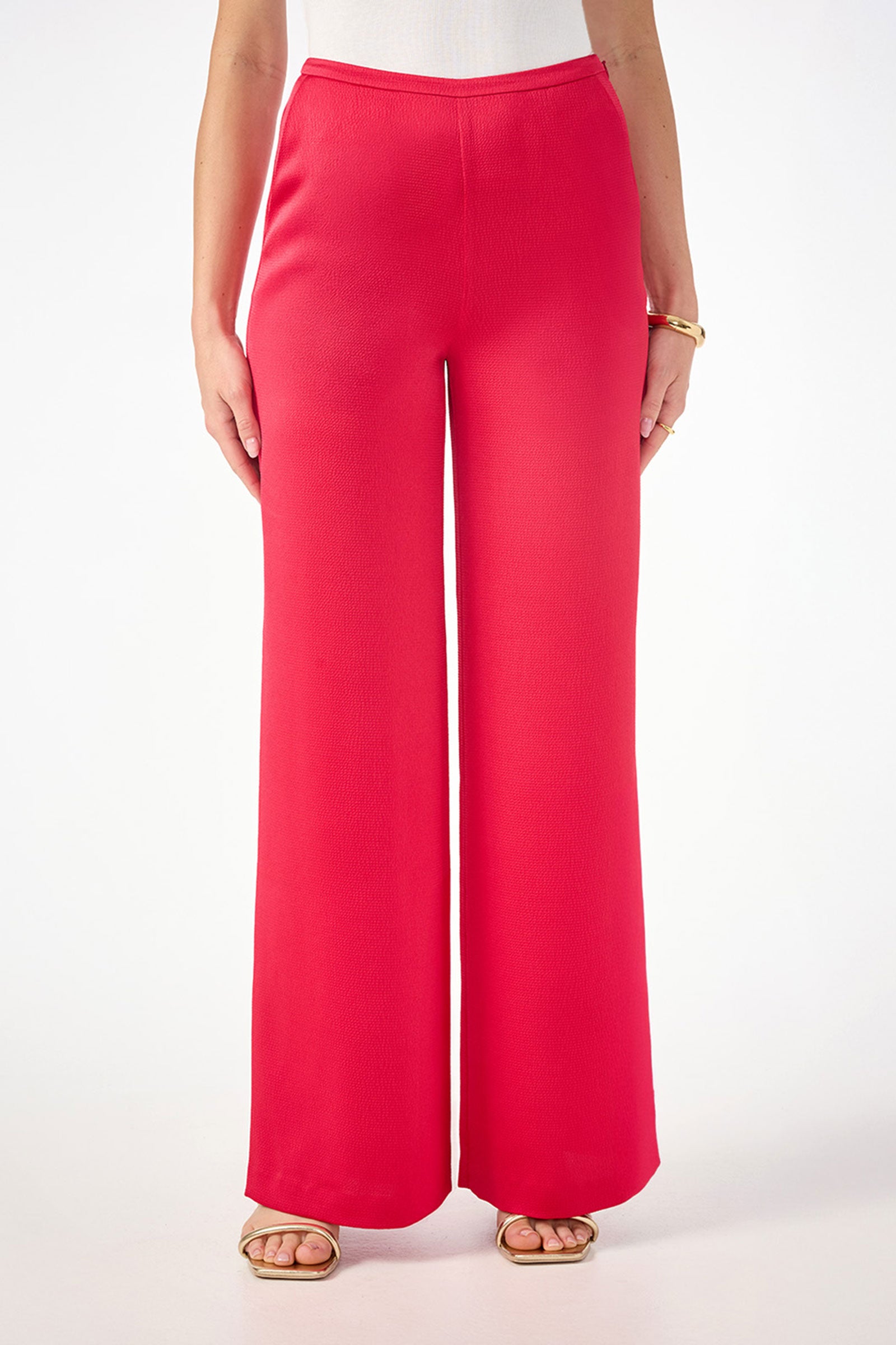 Someone wearing the Tavia Wide-Leg Pant in Cannes Coral (hammered satin), paired with a white top and gold sandals, stands against a plain white background. Only the lower torso and legs are visible.