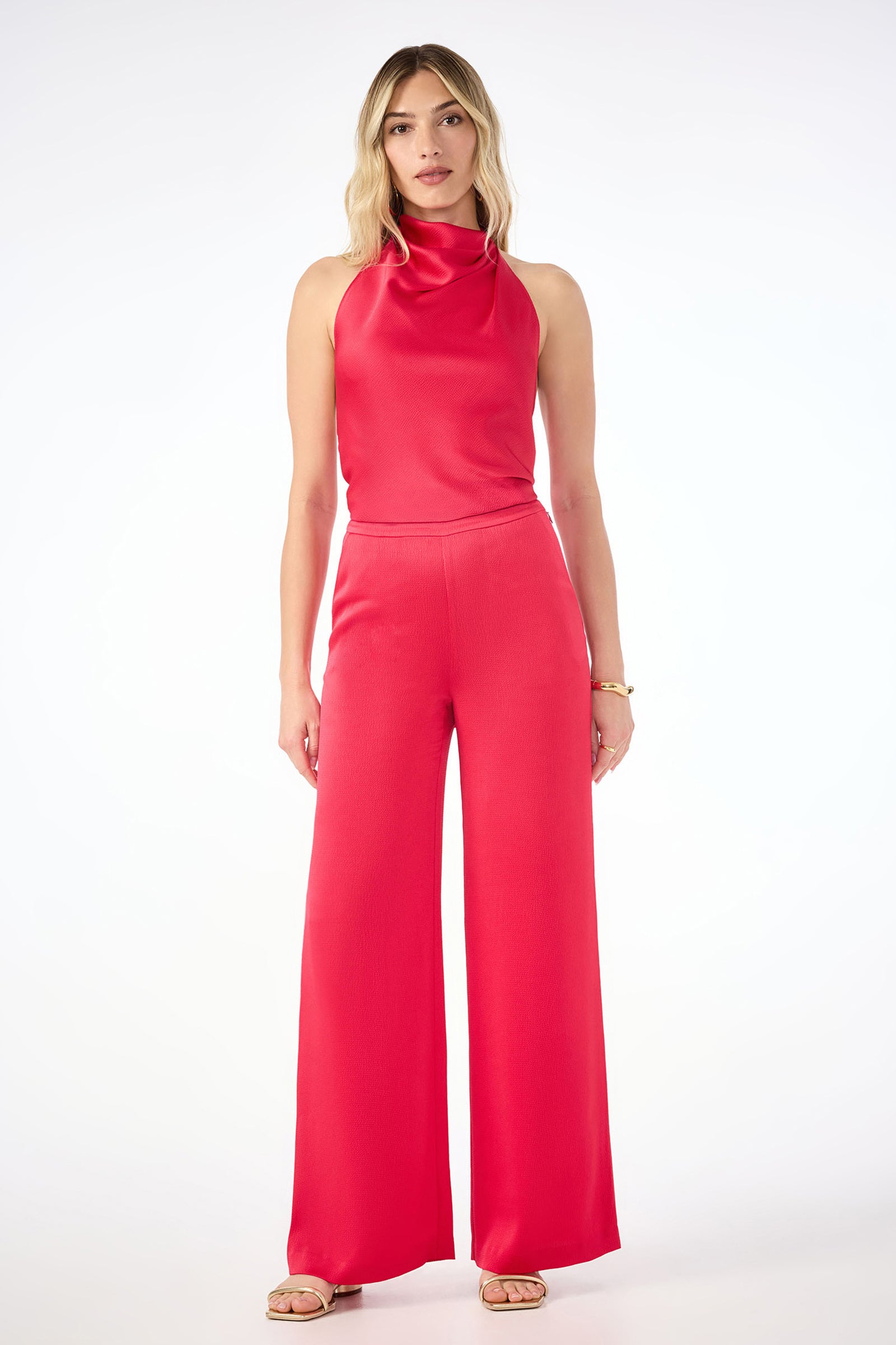 A woman faces forward wearing the Tavia Wide-Leg Pant in hammered satin with a matching sleeveless, high-neck red top. She has long blonde hair and beige sandals, creating a minimal, elegant look against a white background.