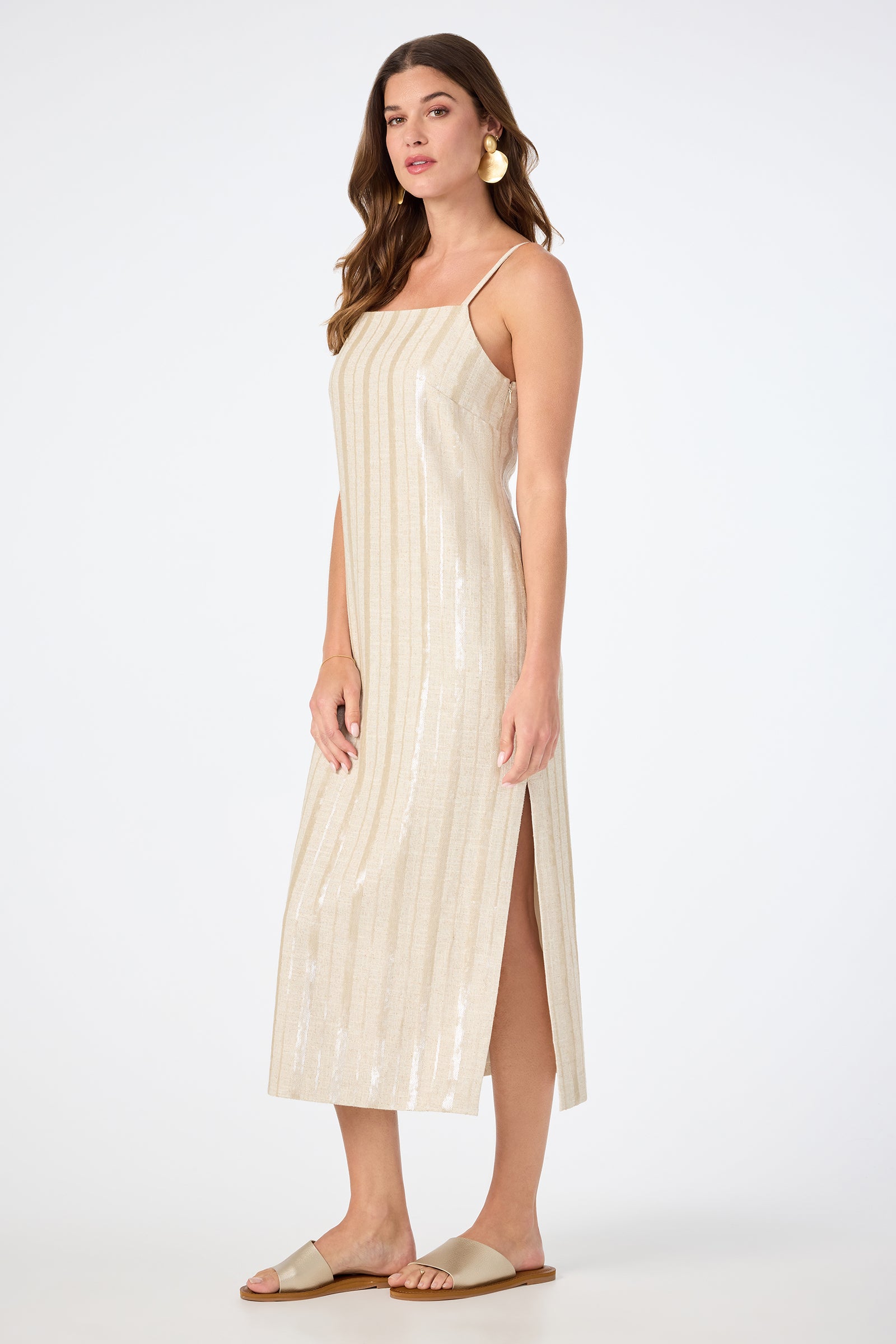A woman wears the Marita 3 Sleeveless Dress—beige, shin-length, with thin straps and side slits—paired with flat beige sandals and gold earrings, standing slightly sideways against a plain white background.