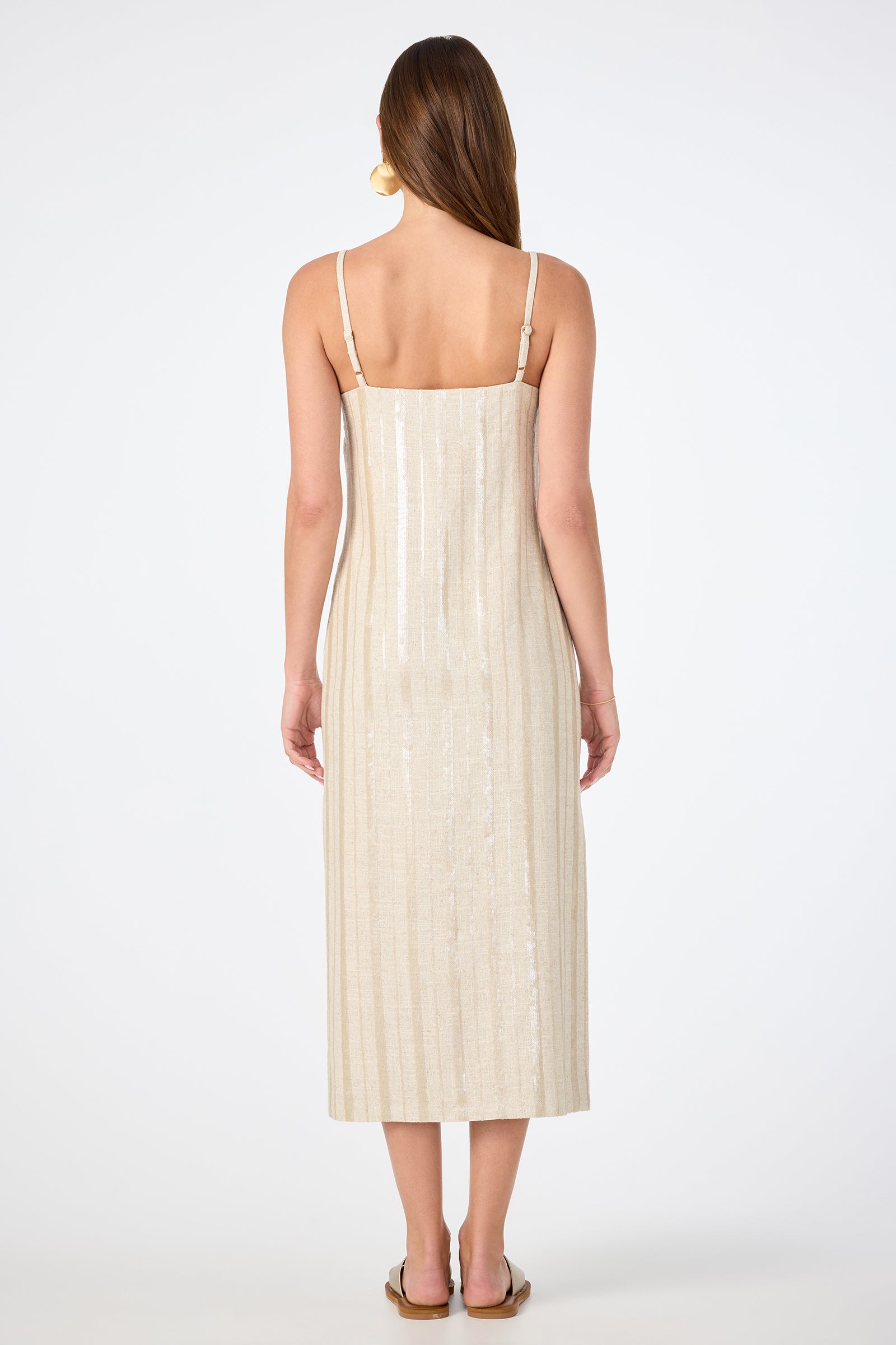 A woman with long straight hair stands with her back to the camera, wearing the Marita 3 Sleeveless Dress in light beige—midi-length with thin straps—and flat sandals. The background is plain white.