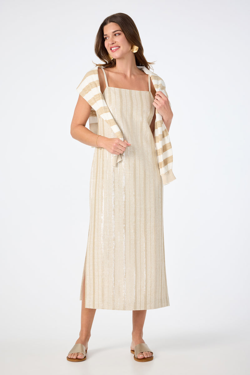 A woman stands smiling against a plain white background, wearing the Marita 3 Sleeveless Dress in Biarritz Beige. The ankle-length dress features subtle vertical stripes. She accessorizes with a striped sweater over her shoulders and flat sandals.