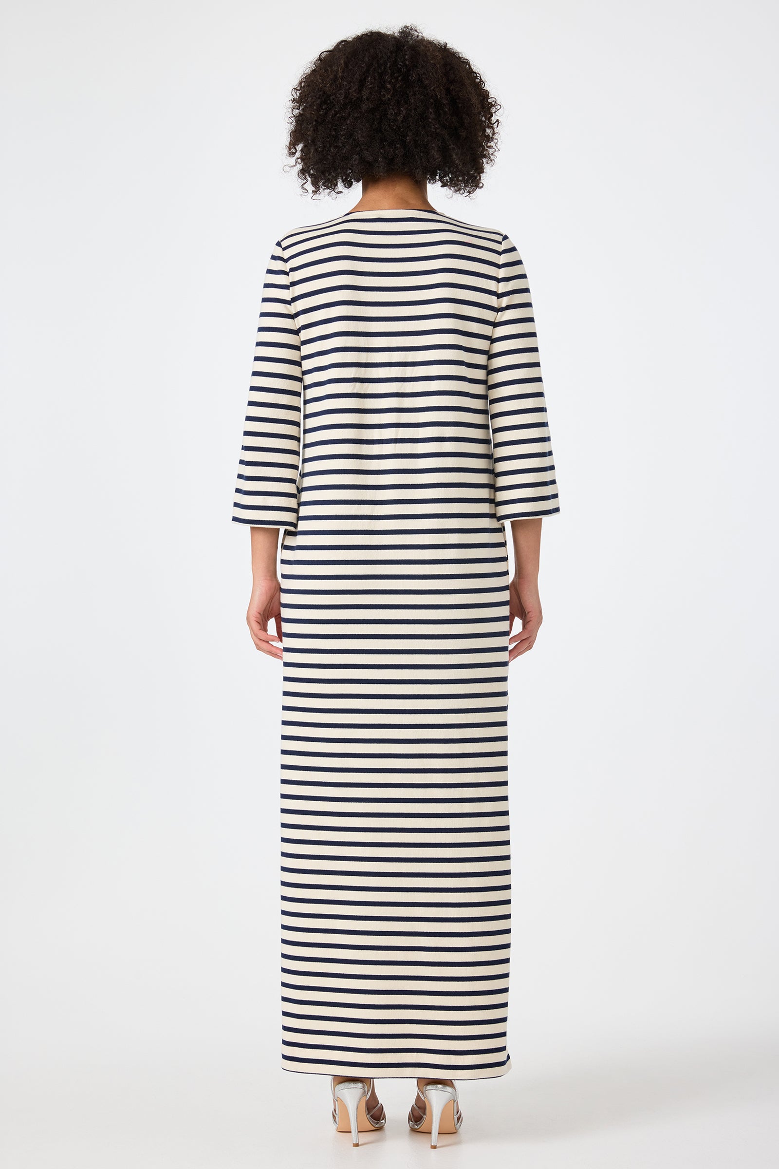 A person with curly hair, seen from behind, wears the Catamaran 3/4 Sleeve Maxi Dress—white with navy horizontal stripes—and heeled sandals, standing against a plain white background.