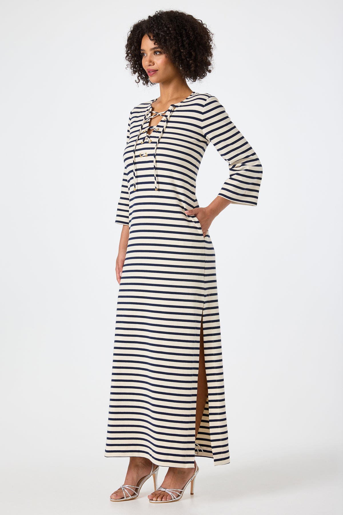 A woman with curly hair stands in profile wearing the Catamaran 3/4 Sleeve Maxi Dress, featuring black and white stripes, a lace-up neckline, side slit, and paired with strappy heeled sandals.