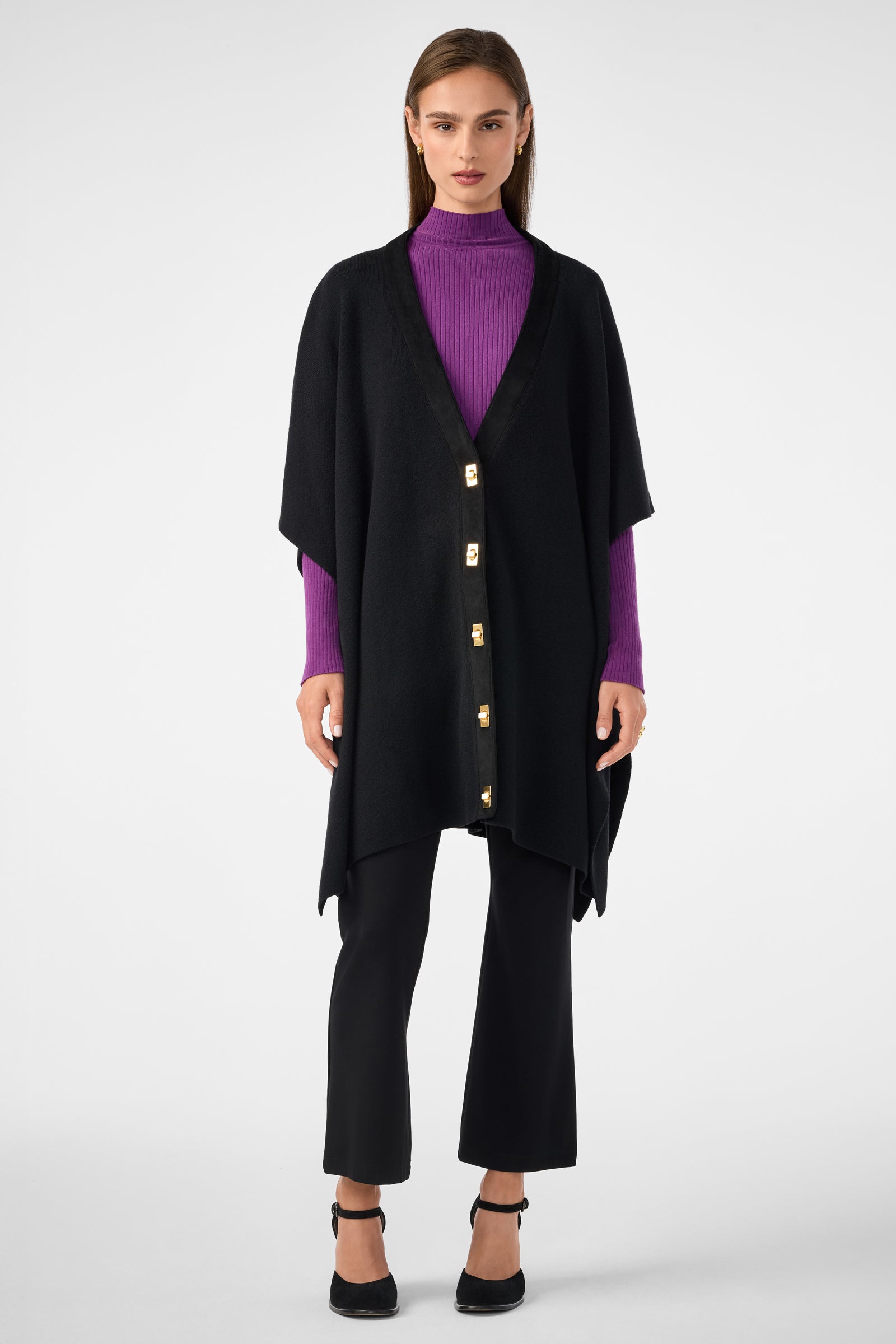 A woman stands against a plain background wearing the Sapphire Poncho over a purple ribbed turtleneck, black cropped pants, and black heeled shoes. She faces forward with a neutral expression.