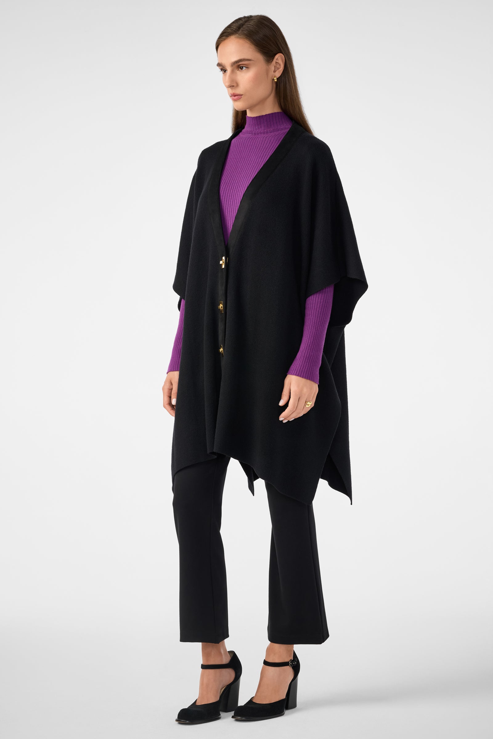 A woman stands against a light gray background wearing the Sapphire Poncho with gold buttons over a purple ribbed turtleneck, paired with black pants and black heeled shoes.