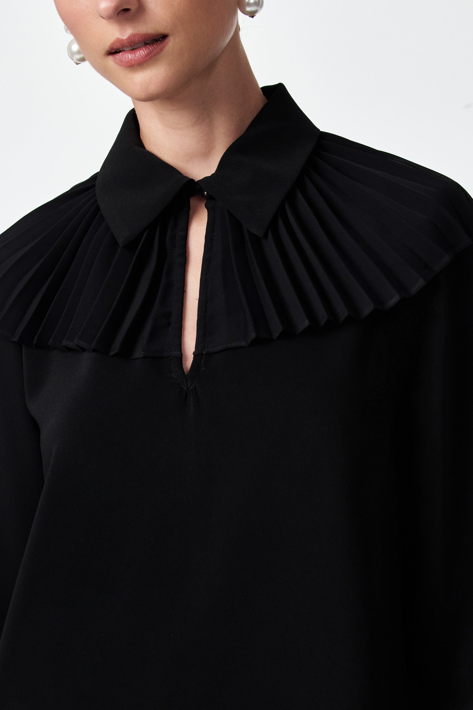 A woman wears the Windmill Top, a black blouse featuring a pleated collar detail and keyhole neckline; her face is partially visible, and she accessorizes with silver hoop earrings.