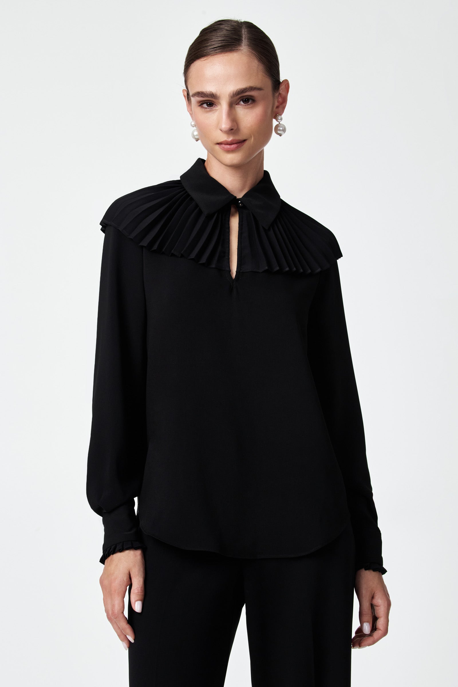 A woman stands against a plain white background wearing the Windmill Top—a long-sleeve black blouse with a pleated cape-like collar—paired with black pants, pearl drop earrings, and her hair pulled back.