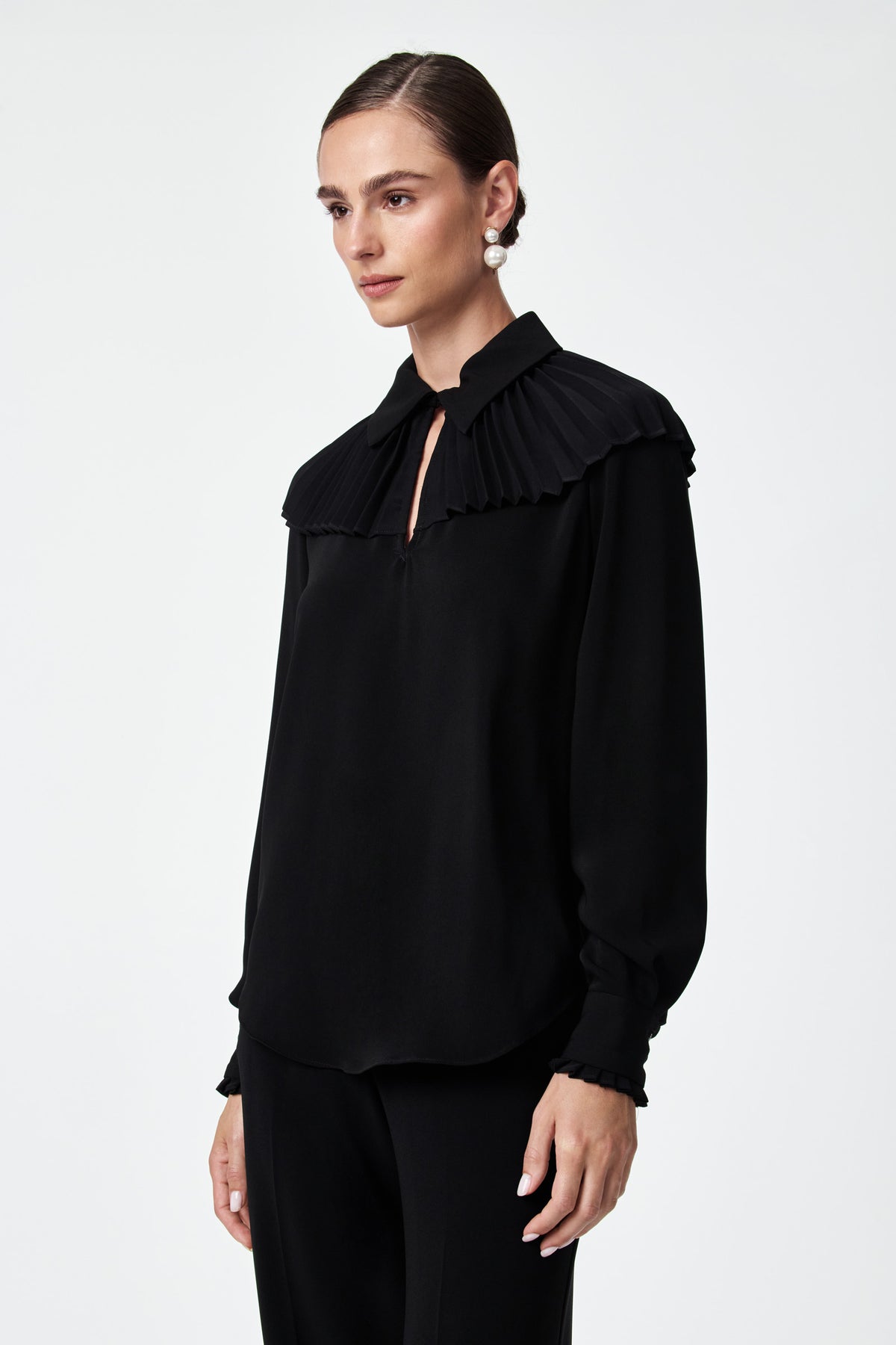 A woman stands against a plain background, wearing the Windmill Top—a black long-sleeve blouse featuring pleated details on the collar and shoulders. She pairs it with black pants, pearl drop earrings, and has her dark hair pulled back.
