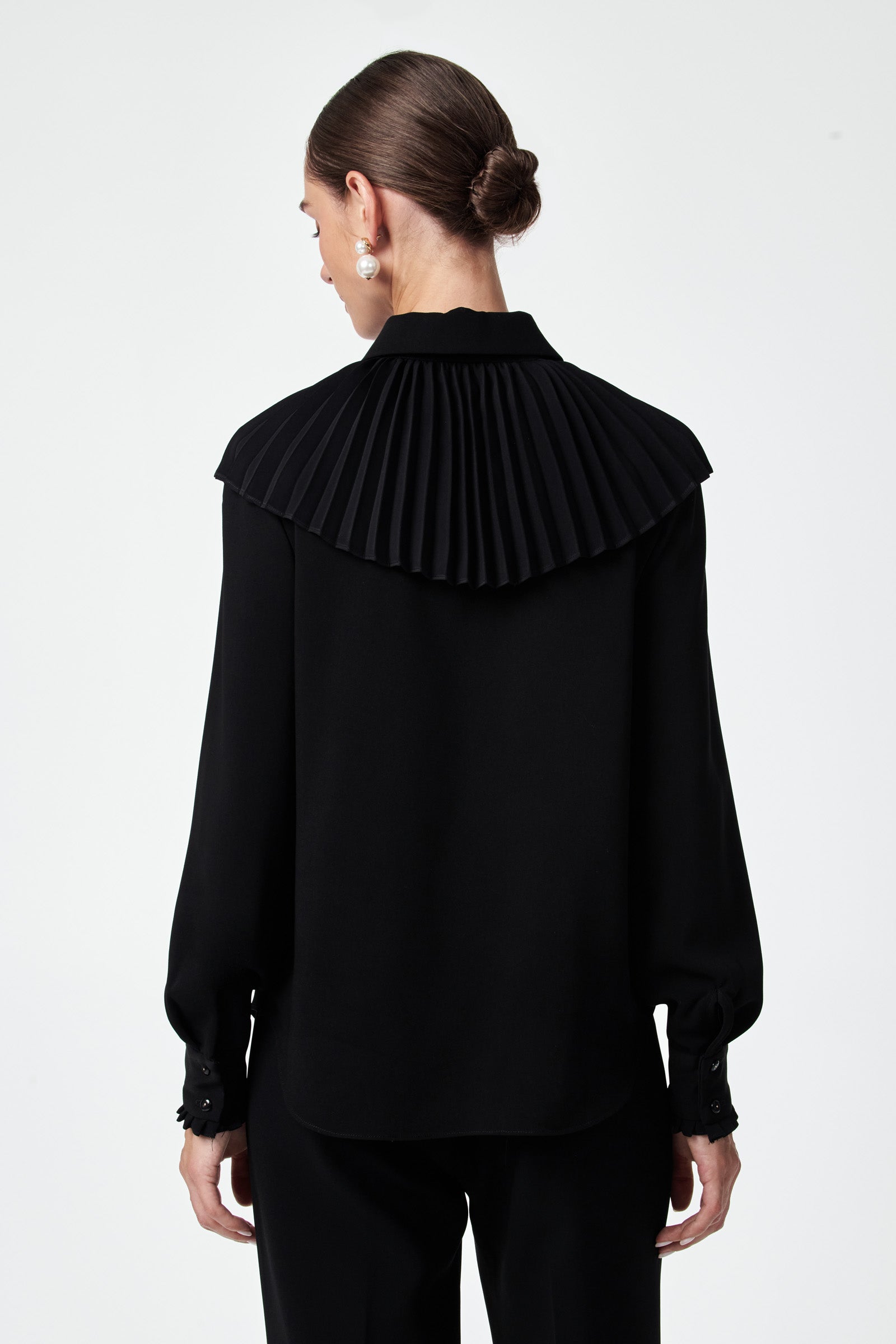 A woman with her hair in a low bun models the Windmill Top, a black long-sleeve blouse featuring a wide, pleated yoke detail on the back, as she faces away from the camera against a plain white background.