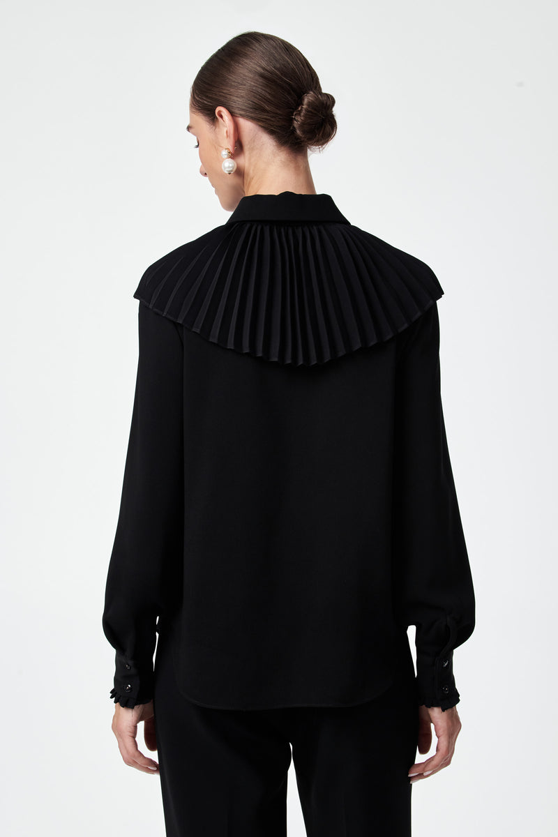 A woman with her hair in a low bun models the Windmill Top, a black long-sleeve blouse featuring a wide, pleated yoke detail on the back, as she faces away from the camera against a plain white background.