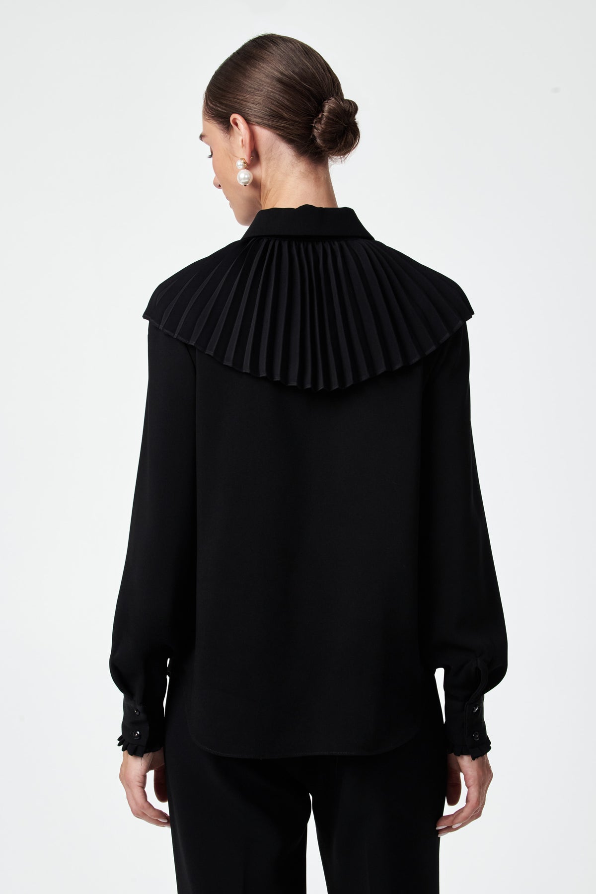A woman with her hair in a low bun models the Windmill Top, a black long-sleeve blouse featuring a wide, pleated yoke detail on the back, as she faces away from the camera against a plain white background.