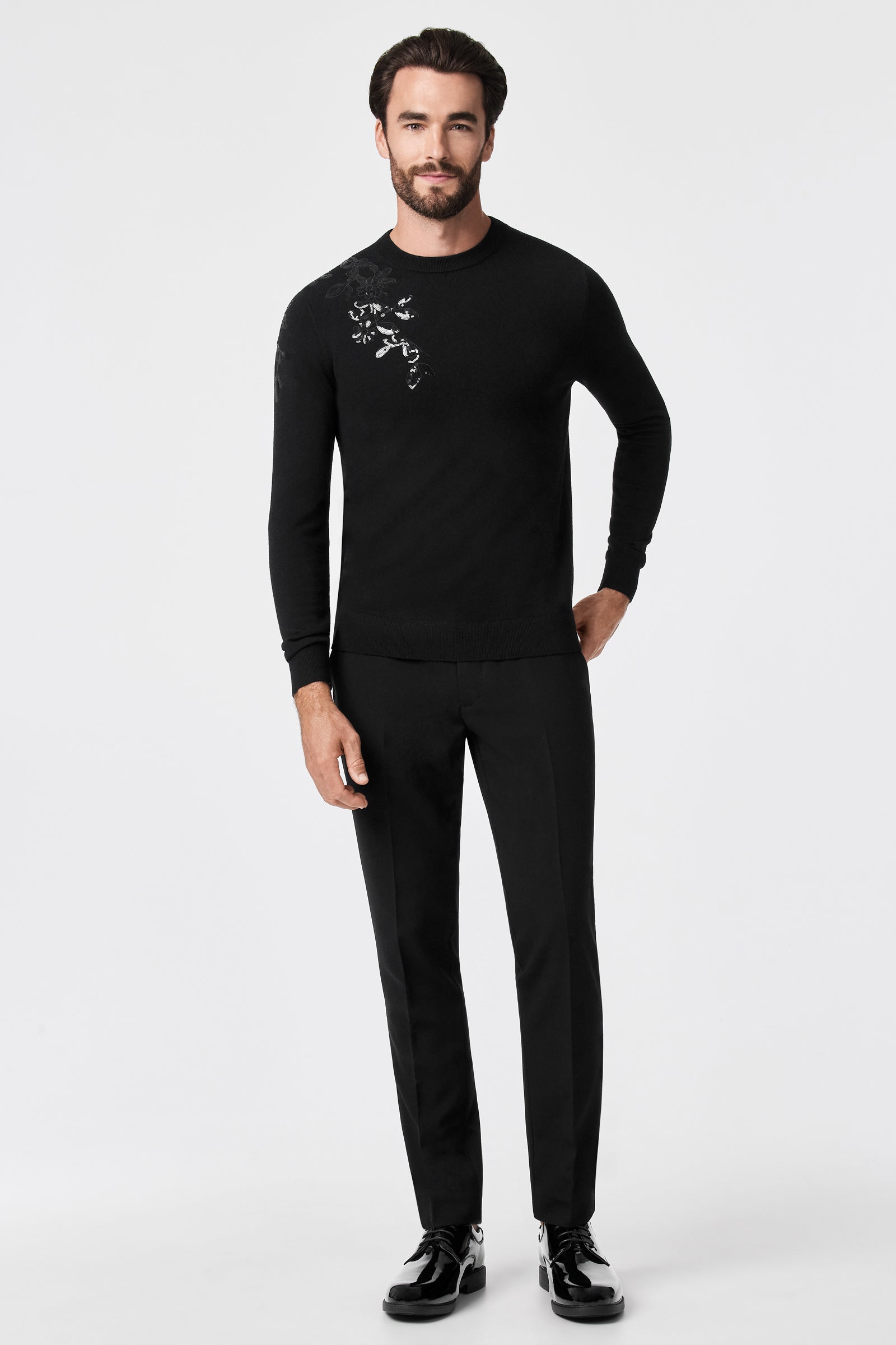 A bearded man wears the Jaxon Sequin Crewneck Sweater—black with a small white floral design on the shoulder—paired with black pants and dress shoes, standing against a plain white background.