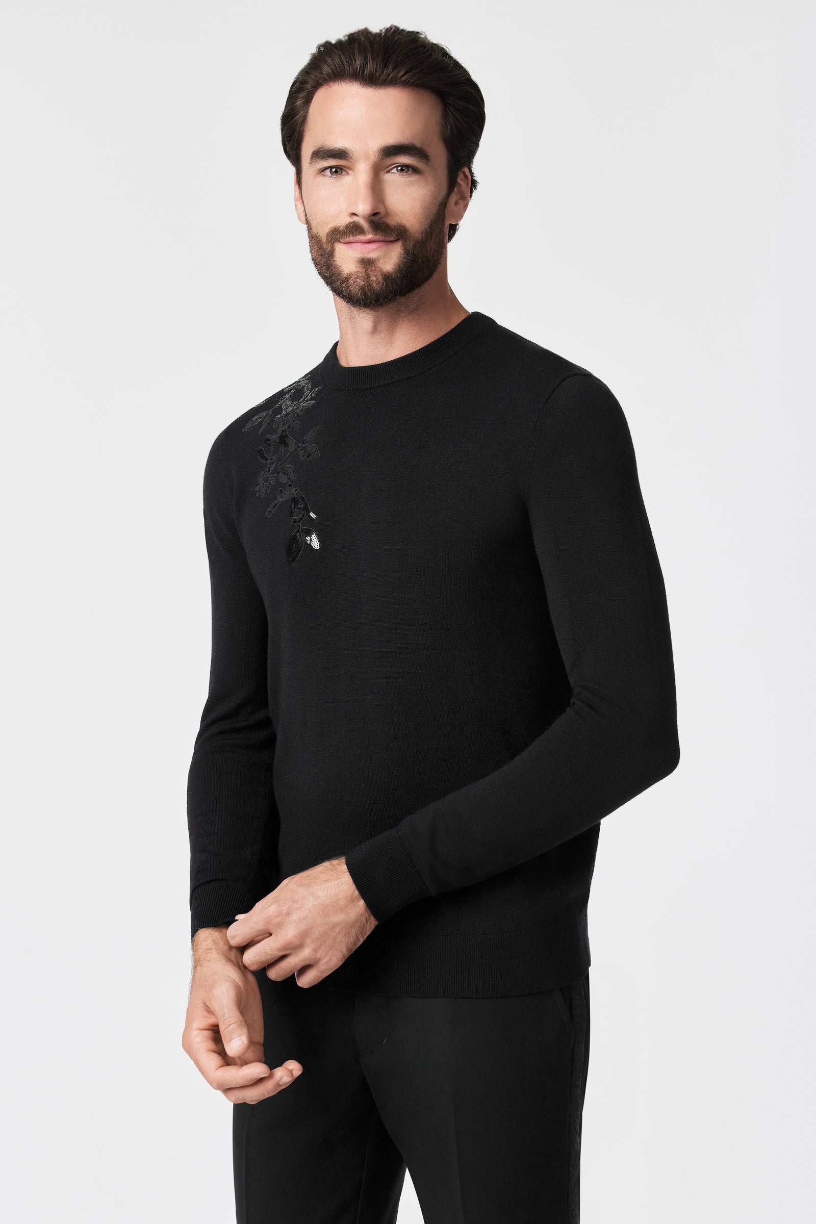 A man with dark hair and a beard models the Jaxon Sequin Crewneck Sweater, a fitted black top featuring a subtle floral design on one shoulder, posed against a plain white background.