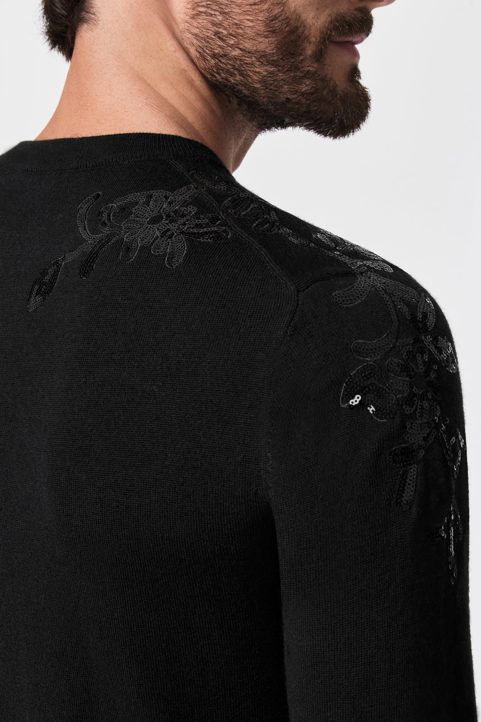 A close-up of a man's upper back and shoulder wearing the Jaxon Sequin Crewneck Sweater, black with subtle embroidered floral patterns near the shoulder, photographed from behind.