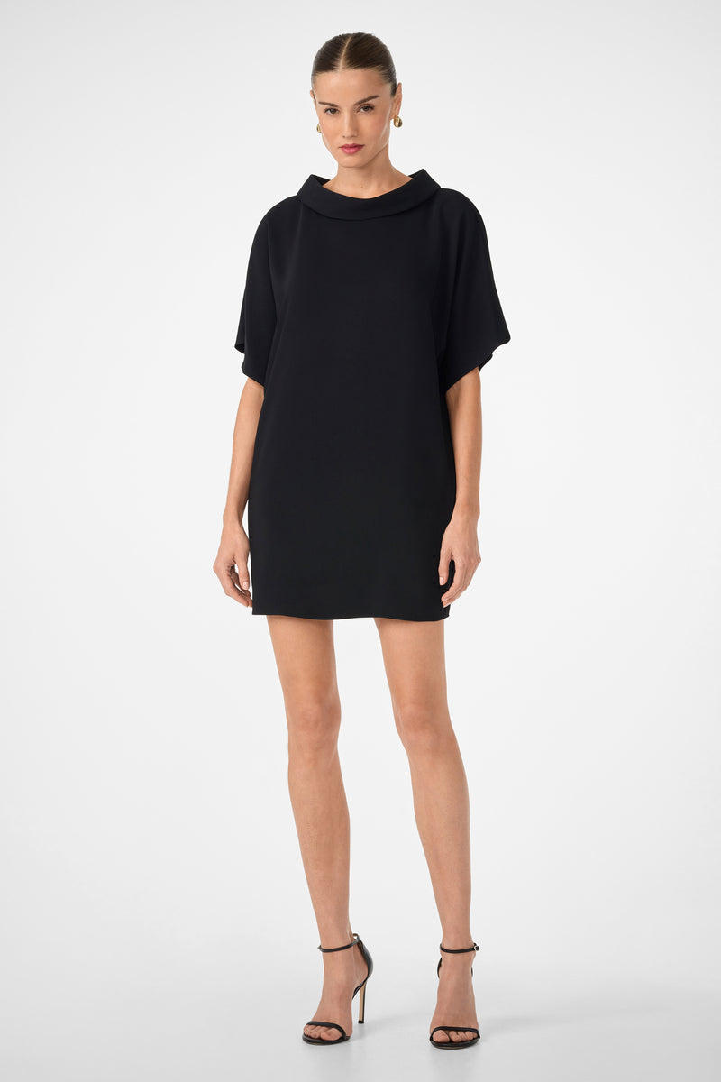 A woman with her hair tied back and a neutral expression stands against a plain light background wearing the black Honeysuckle Short Dolmain Sleeve Shift Dress and black high-heeled sandals.