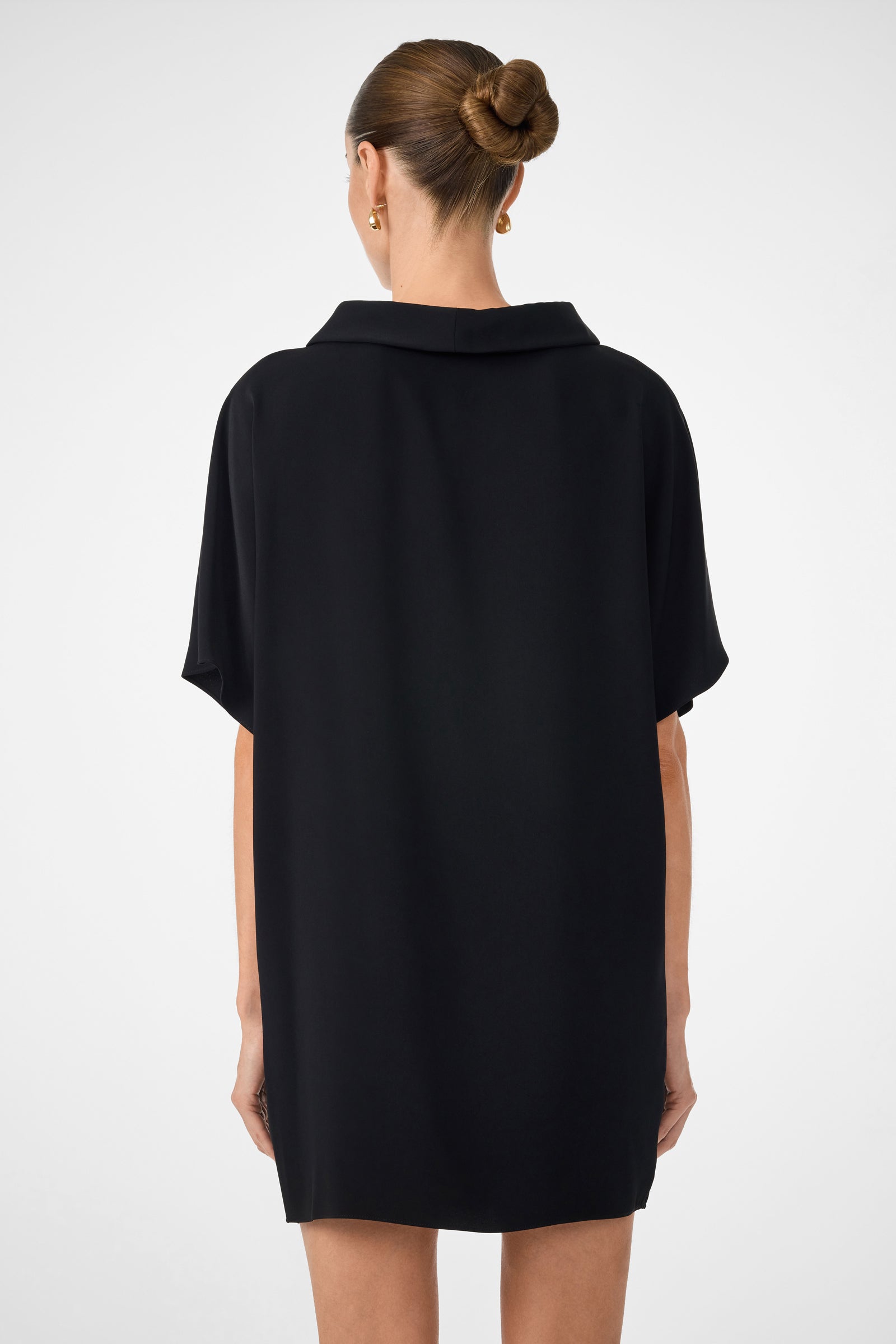 A woman with brown hair in a neat bun wears the black Honeysuckle Short Dolmain Sleeve Shift Dress, facing away from the camera against a plain light background.