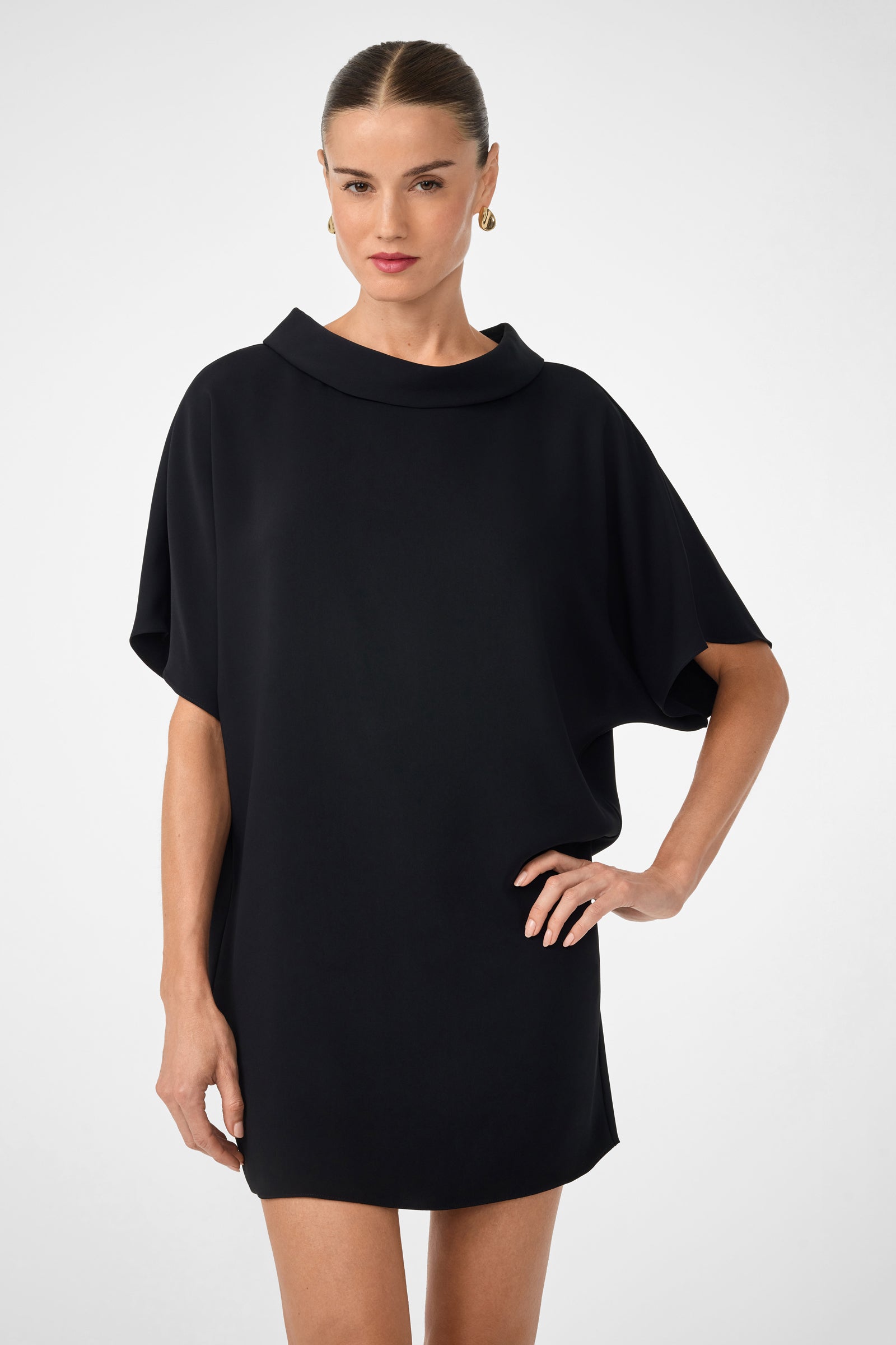 A woman wearing the Honeysuckle Short Dolmain Sleeve Shift Dress, a loose-fitting black dress with short sleeves, stands against a plain background with one hand on her hip, her hair pulled back and gold hoop earrings on.