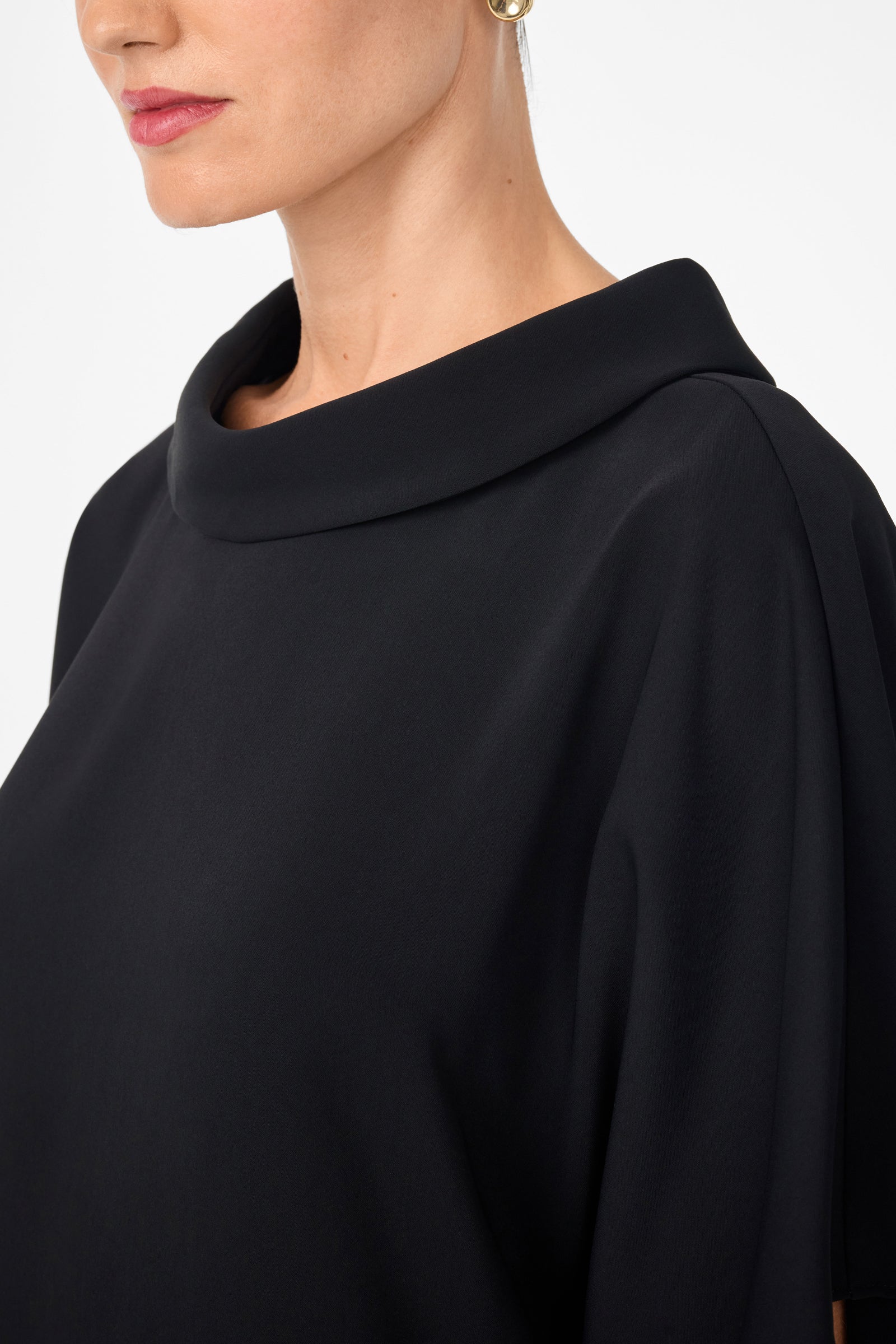 A woman wears the Honeysuckle Short Dolmain Sleeve Shift Dress, a black, high-collar, loose-fitting style with short sleeves. Only her lower face, neck, and upper chest are visible against a white background; she has stud earrings and pink lipstick.