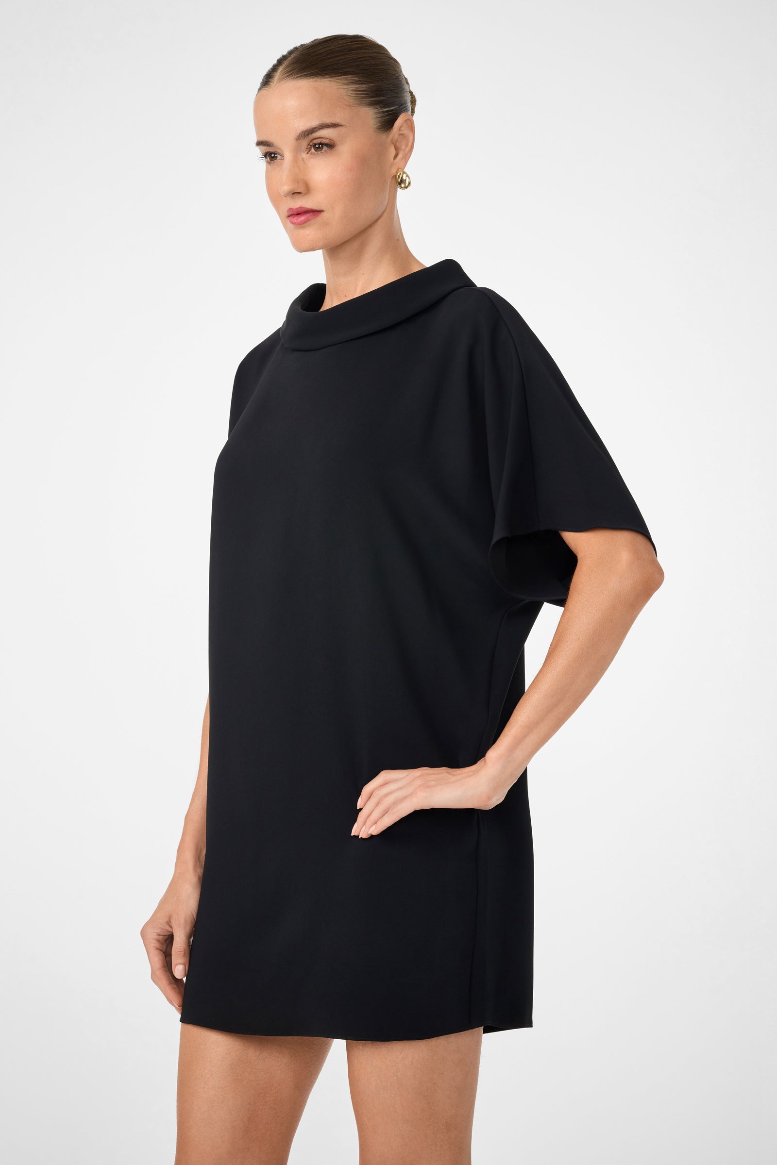 A woman stands against a white background, confidently looking to the side, wearing the Honeysuckle Short Dolmain Sleeve Shift Dress—a short, loose-fitting black dress with short sleeves and a wide, draped neckline. Her hair is pulled back.