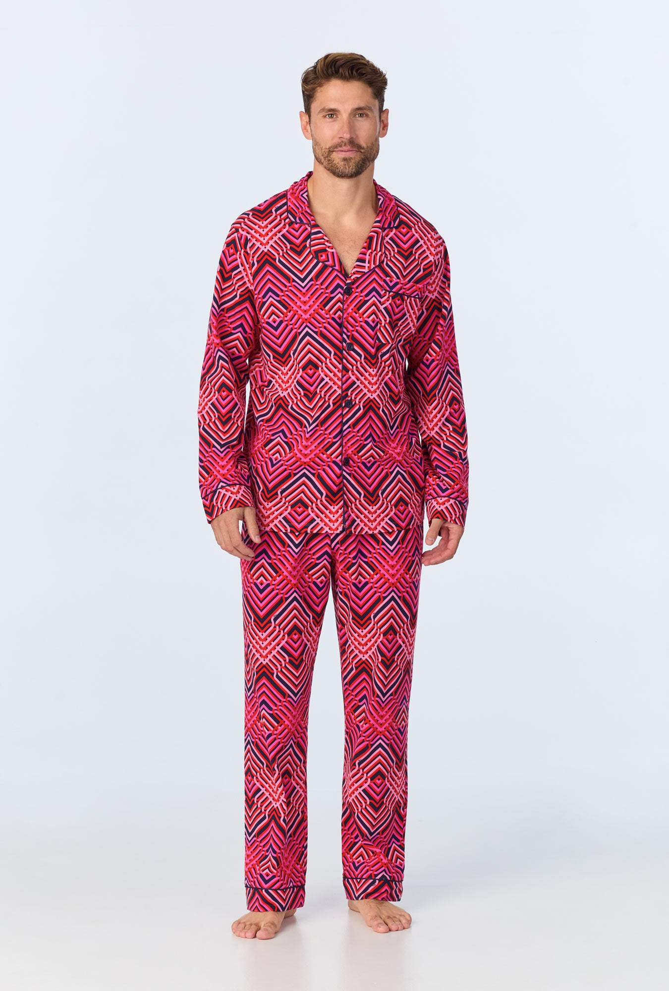 A man stands barefoot against a plain background, wearing the Geo Trellis Mens Long-Sleeve Long Pant Jersey PJ Set in multi, featuring a bold geometric pattern.