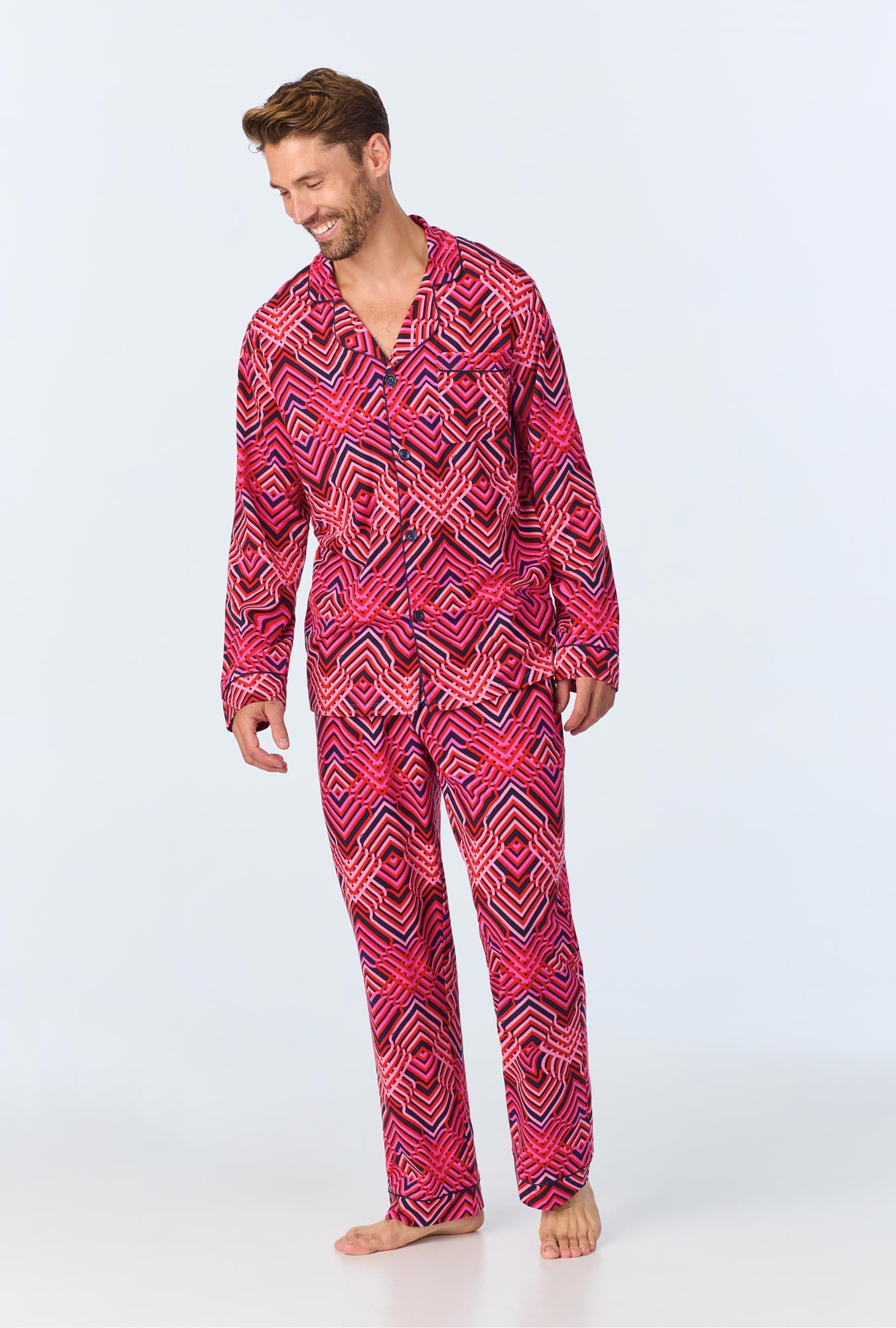A man stands barefoot, smiling and looking down, wearing the Geo Trellis Mens Long-Sleeve Long Pant Jersey PJ Set in red, pink, and black geometric patterns on a light background.