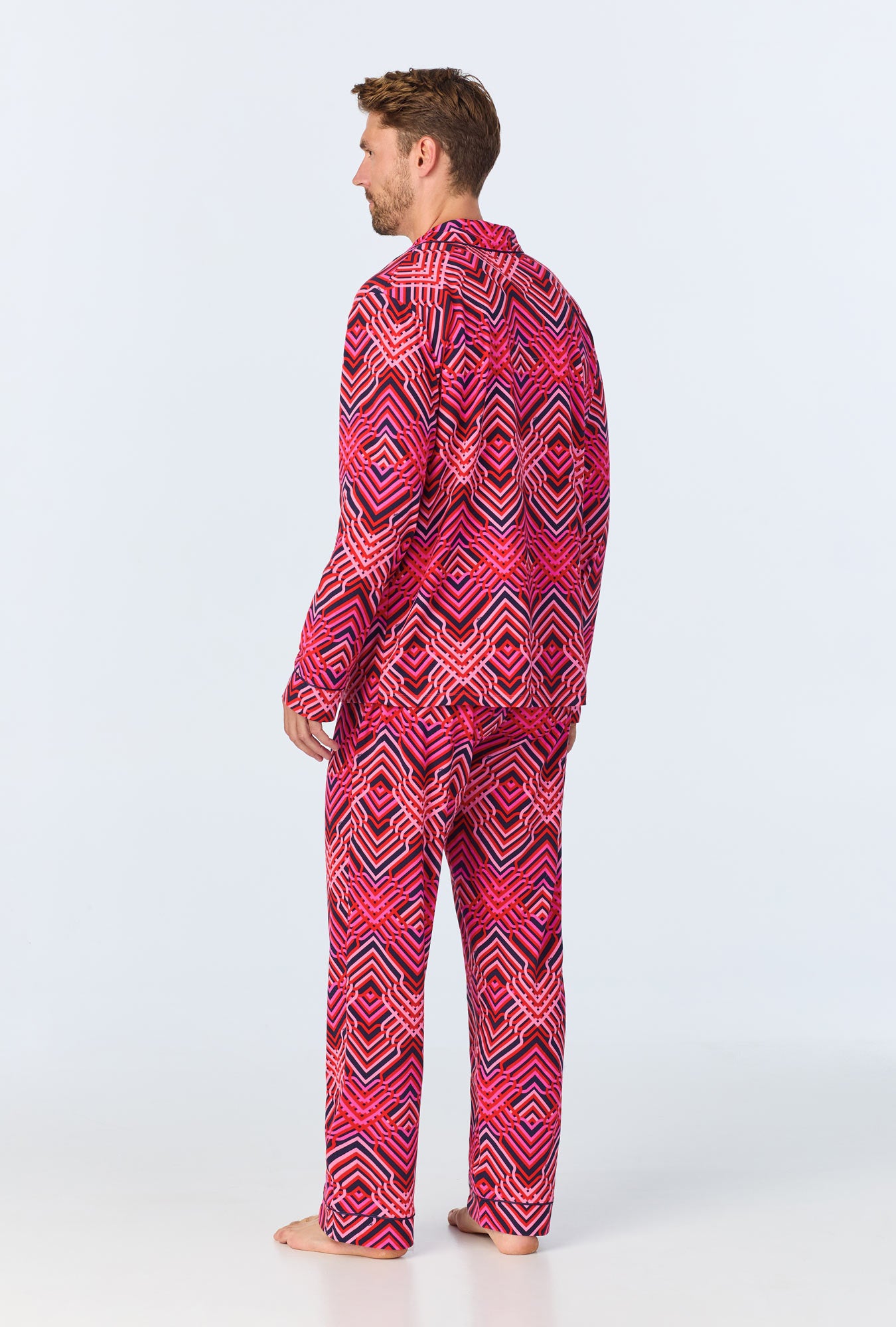 A barefoot man, seen from behind, wears the Geo Trellis Mens Long-Sleeve Long Pant Jersey PJ Set featuring a bold geometric pattern in red, pink, black, and white against a plain light background.