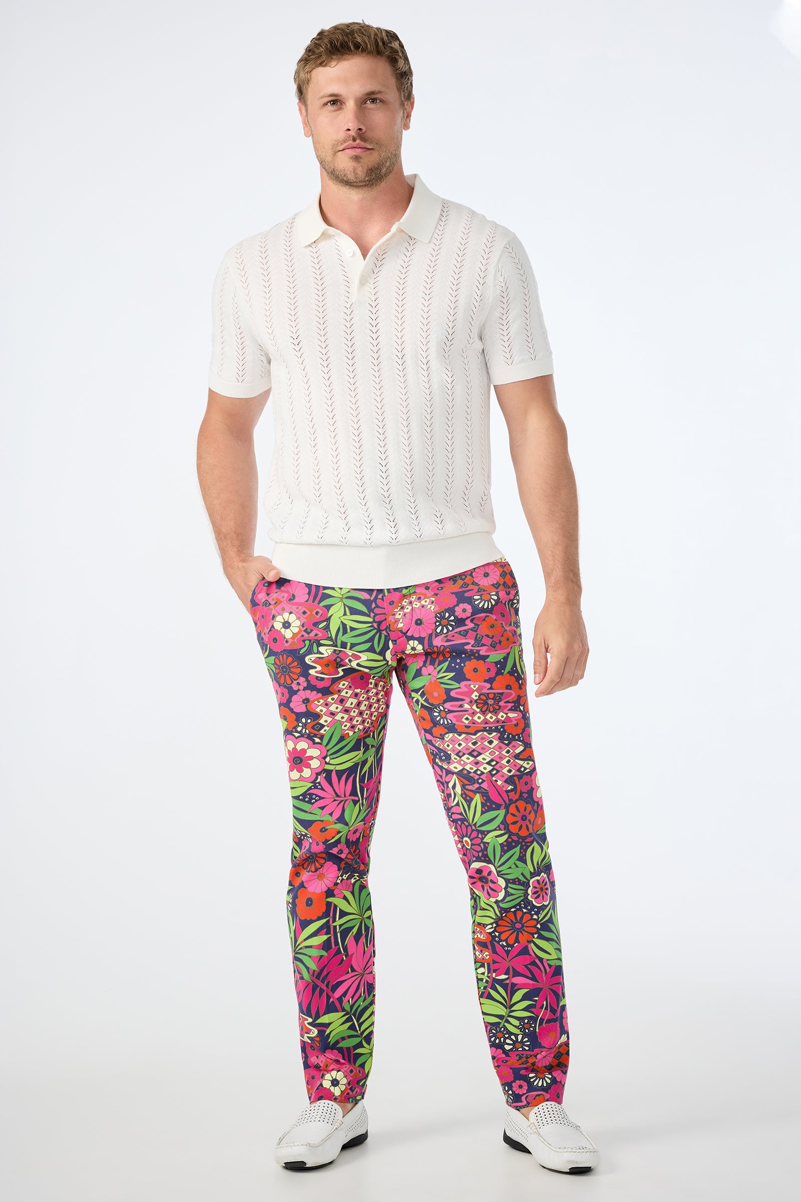 A man stands against a white background, wearing a white short-sleeve knit polo shirt, white shoes, and the Clyde Slim Trouser - Fumi Floral featuring vibrant red, pink, and green patterns.
