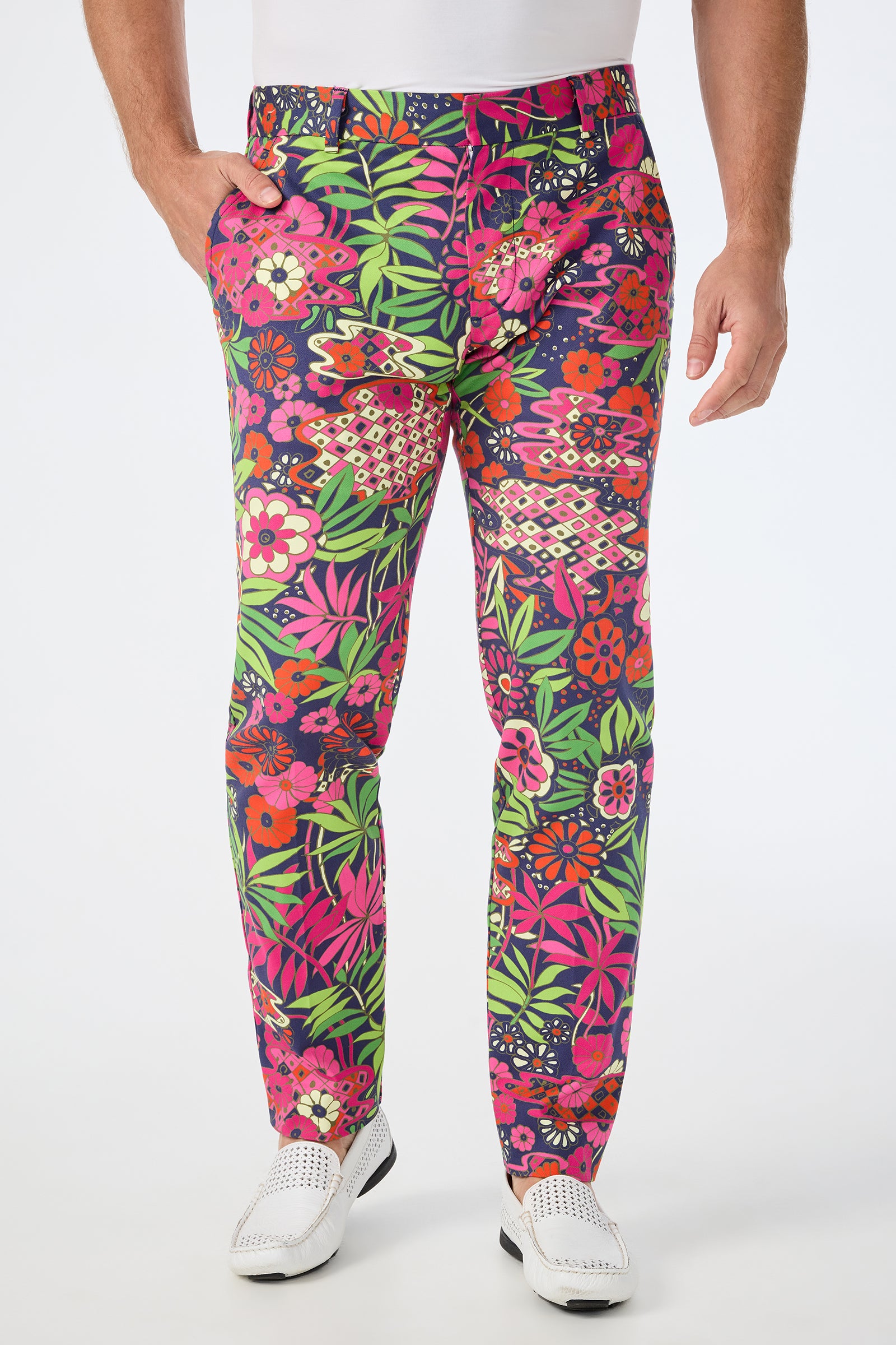 A man wearing the Clyde Slim Trouser in Fumi Floral, featuring vibrant green, pink, red, and purple flowers, paired with a white shirt, belt, and shoes. He is shown from shoulders to feet against a plain background.