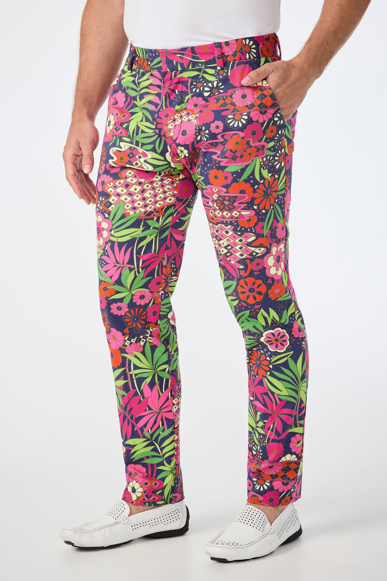 A man wears the Clyde Slim Trouser - Fumi Floral, featuring a pink, red, green, and yellow floral print, styled with a white shirt and white perforated shoes against a plain white background.
