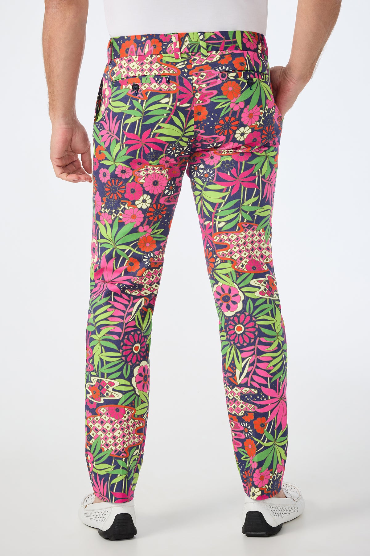 A person stands facing away, wearing the Clyde Slim Trouser - Fumi Floral, featuring a vibrant pink, green, orange, red, and purple floral and geometric print, paired with white shoes and a white shirt.