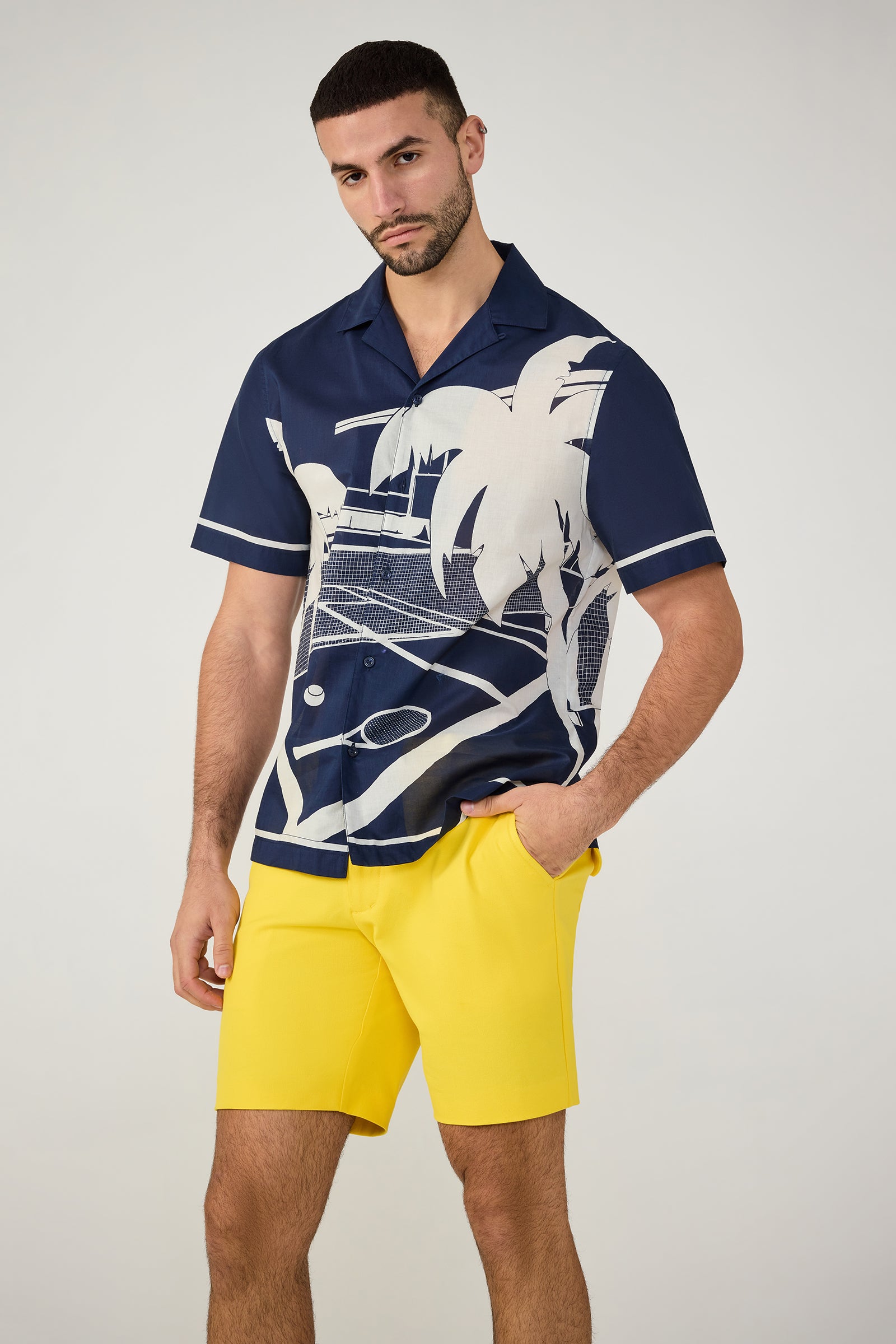 A man with short dark hair, a beard, and a neutral expression wears the Josue Short-Sleeve Shirt in Ink/Whitewash Tennis Print, paired with bright yellow shorts. He stands against a plain background with one hand in his pocket.
