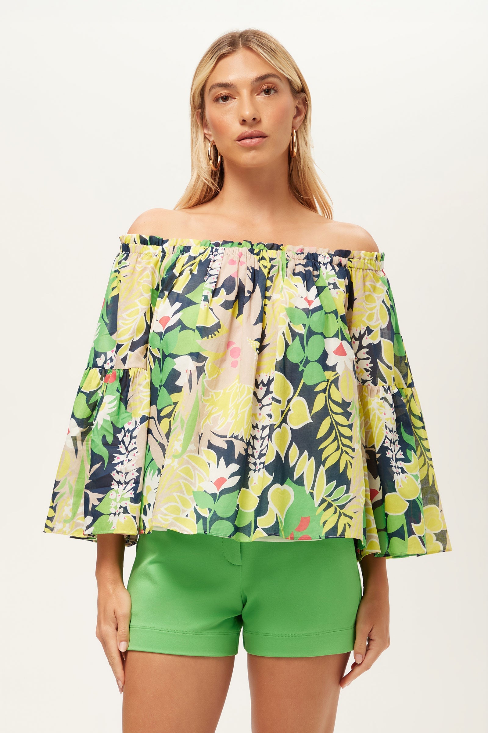 Woman wearing the Perry Off-Shoulder Top - PS Eternal in Palm Springs Eternal, featuring wide sleeves and a floral pattern, paired with bright green shorts against a plain light background.