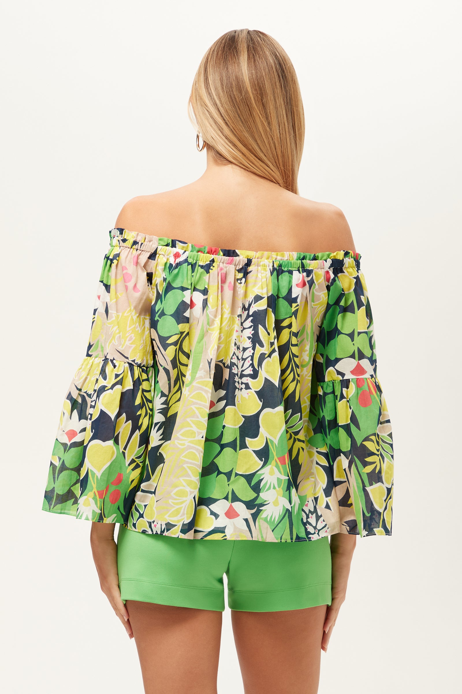 A woman with long, straight hair is seen from behind wearing the Perry Off-Shoulder Top - PS Eternal, a loose floral top in green, yellow, pink, and white, paired with bright green shorts against a plain background.