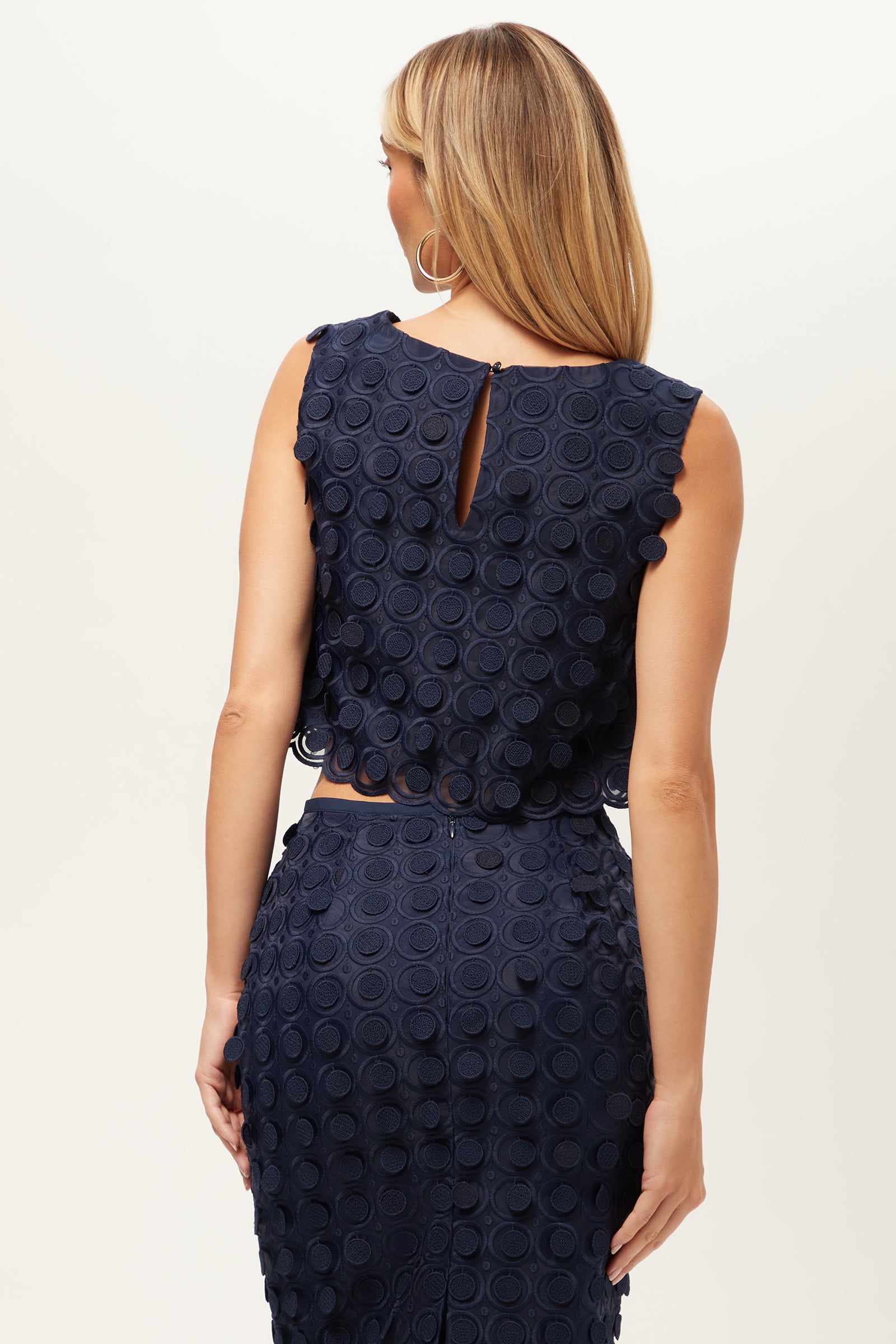 A woman with long blonde hair wears the Serve Sleeveless Top in Agassi Embroidery, featuring navy blue textured fabric with circular patterns, viewed from the back against a plain light background.