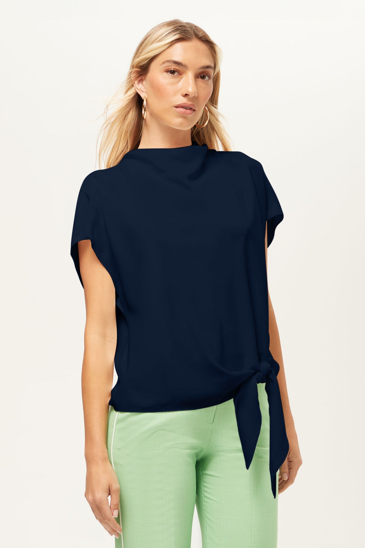 A woman with long blonde hair stands facing forward against a plain, light background, wearing the Bisque Top—a navy short-sleeve top with a side tie—and light green pants.
