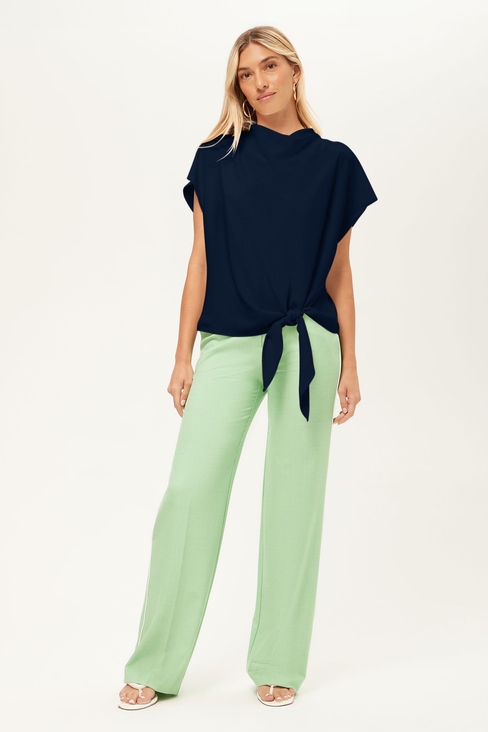 A woman with long blonde hair stands against a plain background, wearing the Bisque Top— a short-sleeved navy top with a knotted waist—paired with light green wide-leg pants and open-toed heels.